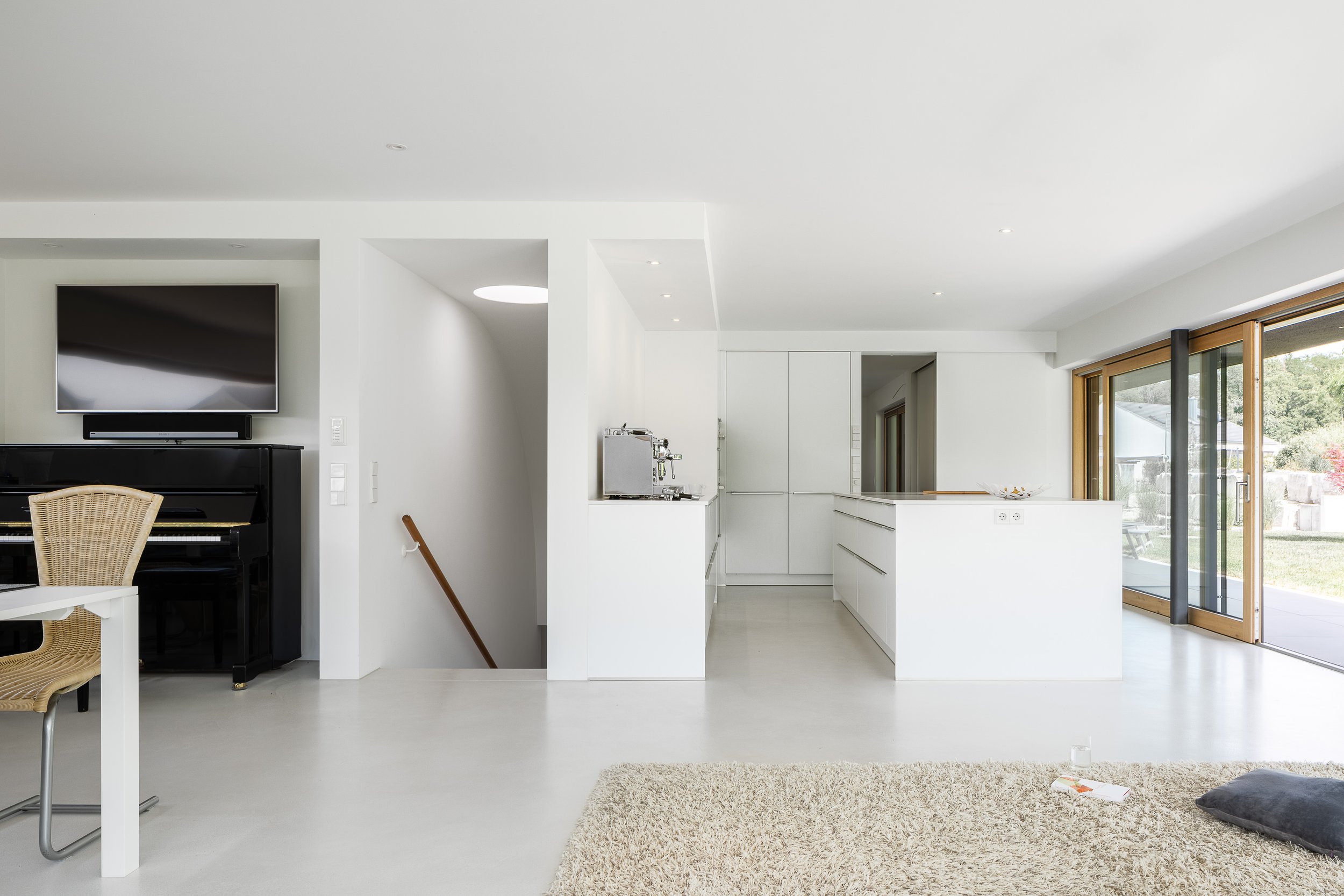 Black Box Haus, Bruchsal, von Henecka Architekten BDA, Offene, helle moderne Wohnzimmerküche mit großen Fenstern, weißen Möbeln, einem schwarzen Klavier, einem Esstisch mit Stühlen und einem weichen Teppich.