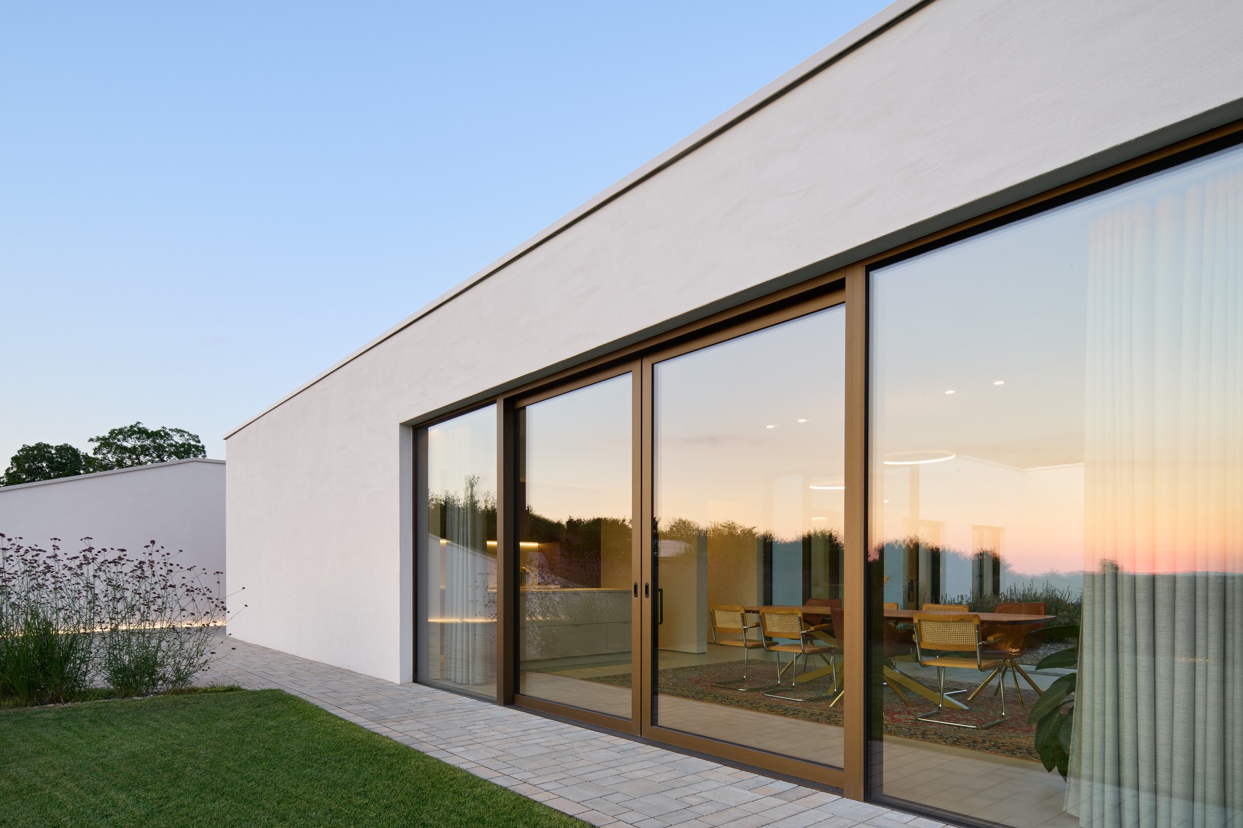 Haus im Weinberg, Bruchsal, von Henecka Architekten BDA, Moderne weiße Hausfassade mit großen Glasschiebetüren und Blick ins Innere, Abendlicht, minimalistische Architektur, Garten mit Rasenflächen und Pflanzen.