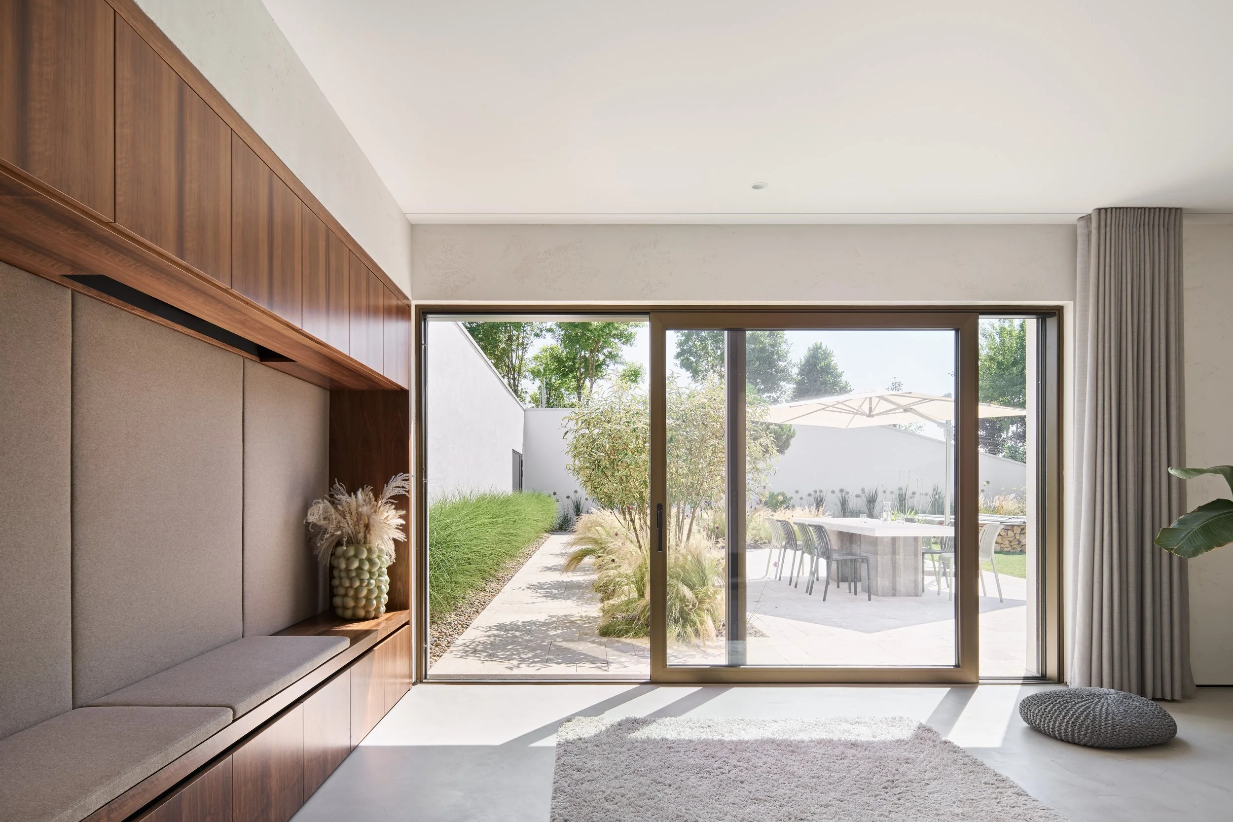 Innenraum eines modernen Wohnzimmers mit großen Glas-Schiebetüren, Blick auf einen Garten mit Sitzgruppe im Freien, Gardinen an der Seite, und eine große, bequeme Sitzbank an der linken Seite.