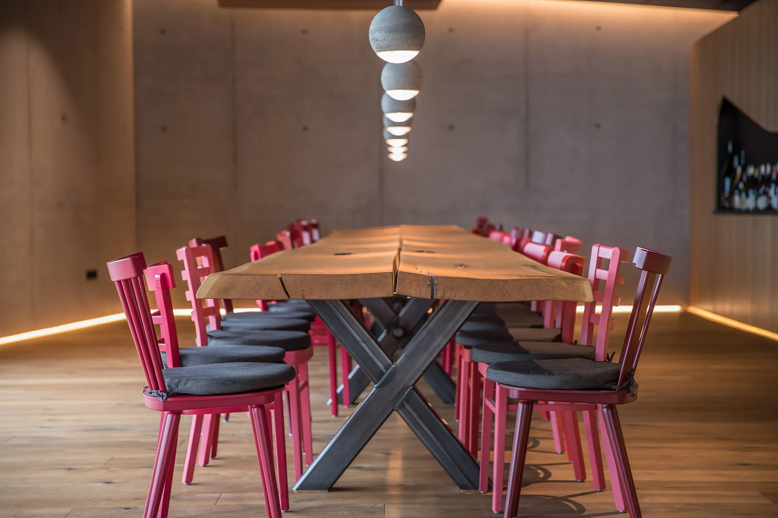 Ein langer Holztisch mit roten Stühlen in einem modernen Raum mit hölzernen Wänden und Decke, beleuchtet von einer Reihe von runden, grauen Pendelleuchten.