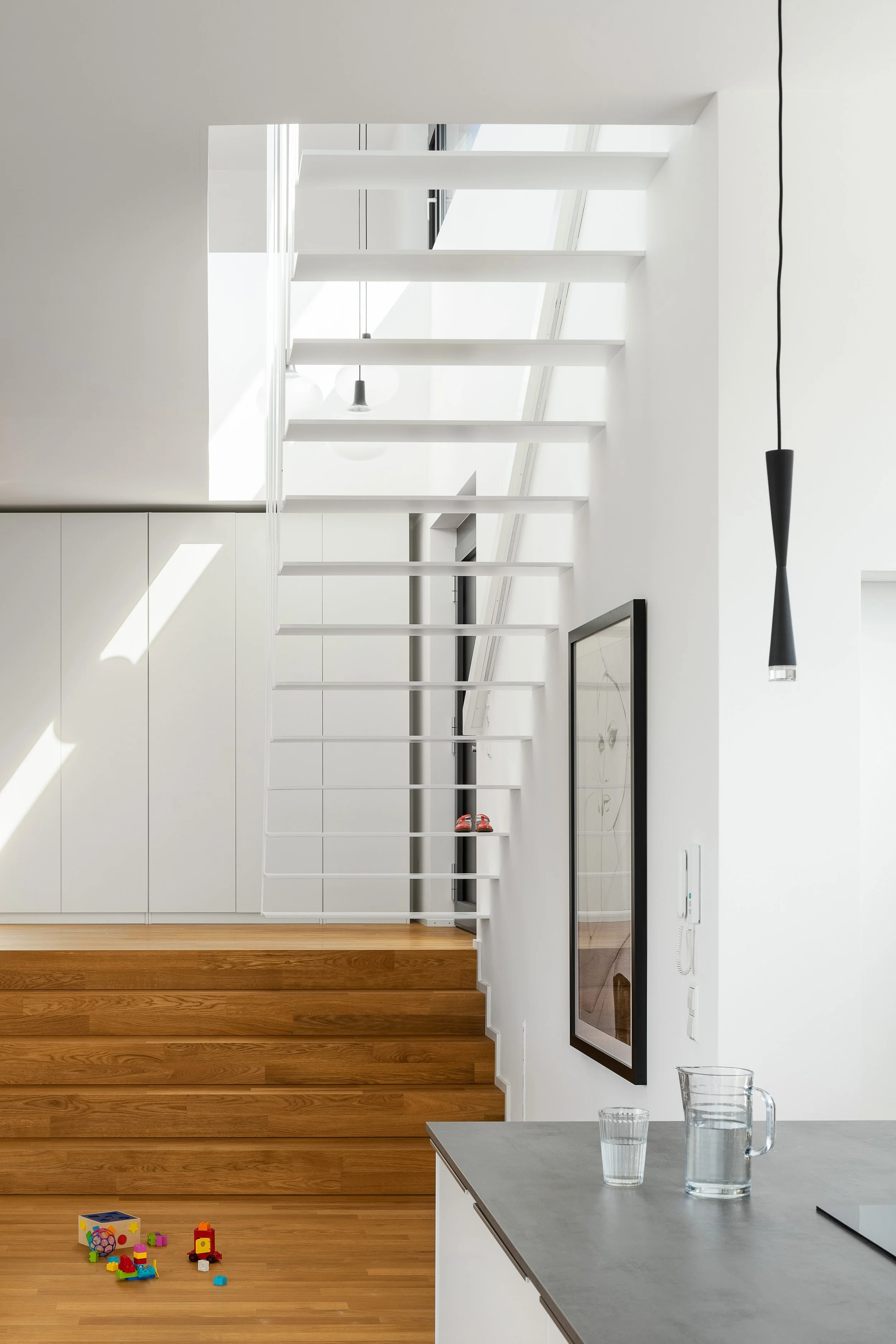 Split Stair Haus Bruchsal von Henecka Architekten BDA Küche mit weißen Wänden und modernen Möbeln. Eine Treppe mit Holzstufen führt nach oben, während eine große schwarze Bilderrahmenwanddekoration an der Wand hängt.