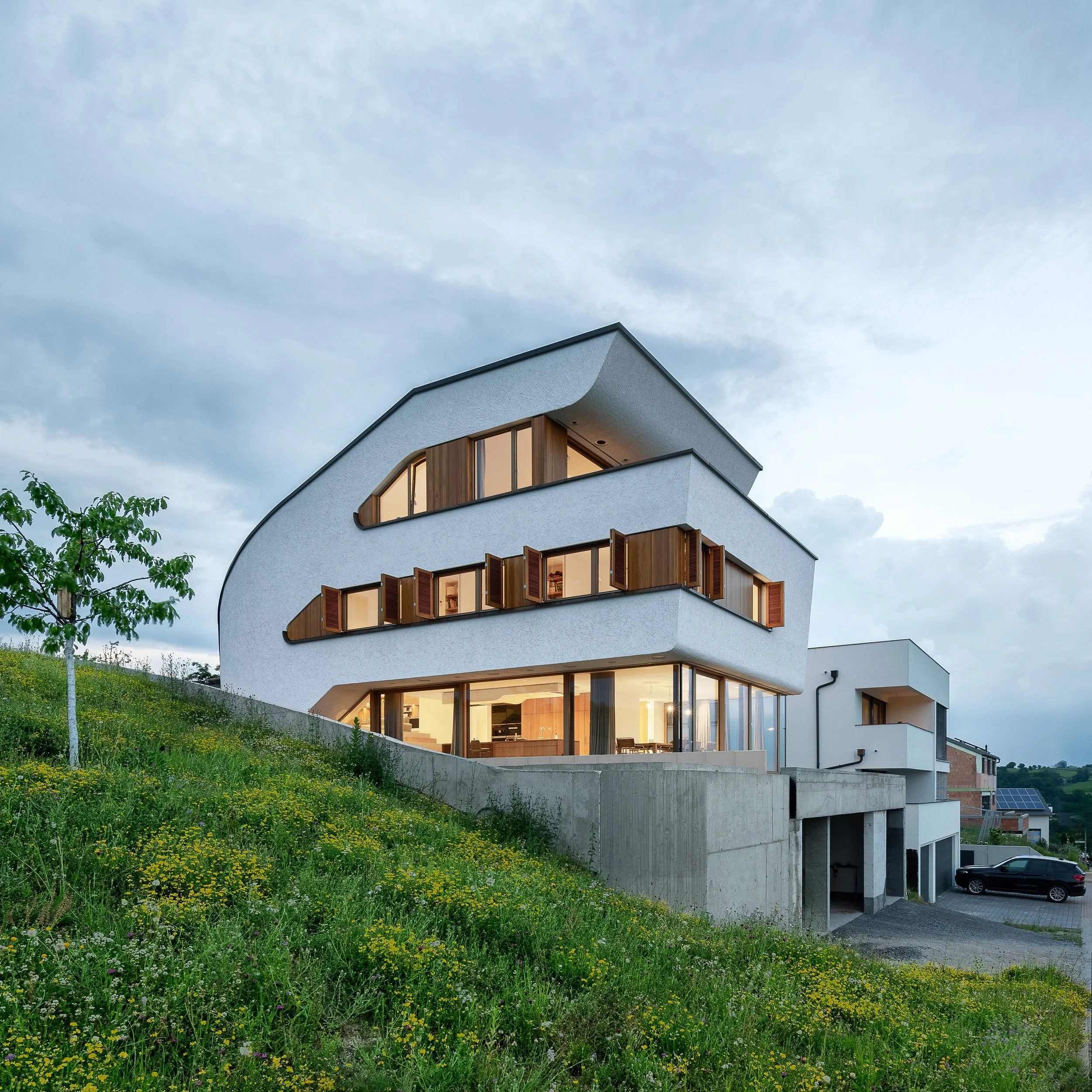 Modernes weißes Mehrfamilienhaus mit Holzelementen und großen Fensterflächen, umgeben von einem grünen Hang mit Wildblumen, unter einem bewölkten Himmel.