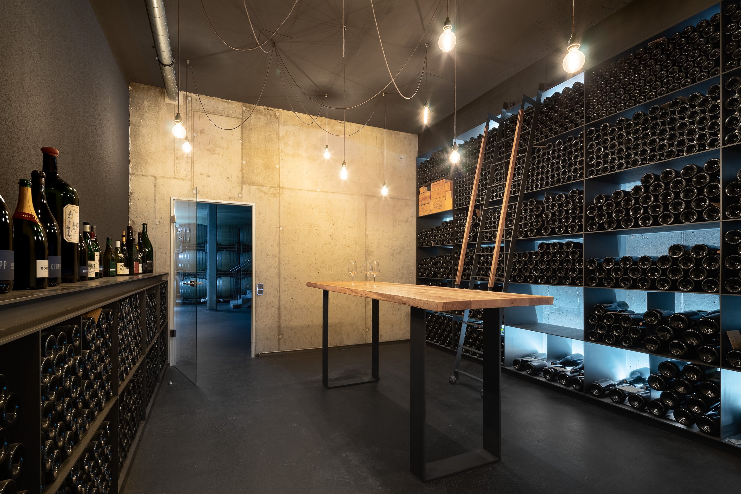Vinothek Weingut Klumpp Bruchsal von Henecka Architekten BDA, Ein Weinkeller mit dunklen Weinflaschen in Stahl-Regalen, einem Holztisch mit zwei Weingläsern und einer Beleuchtung durch Glühbirnen.