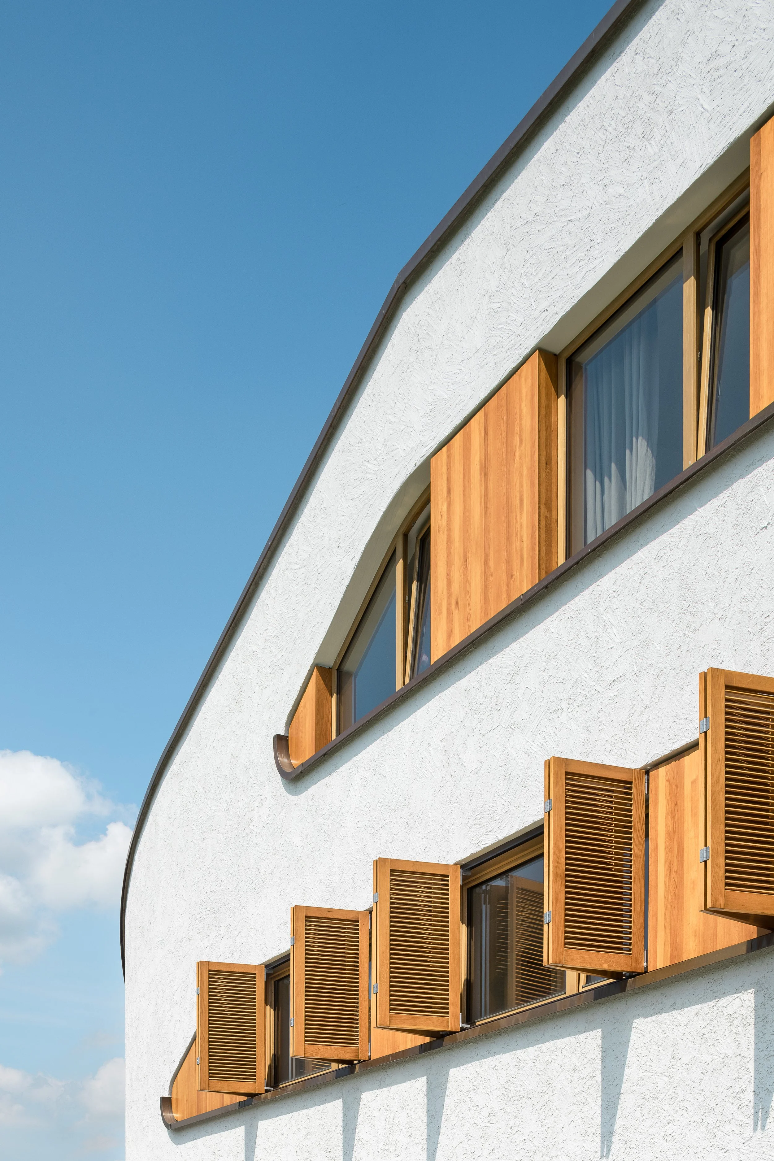 Ein modernes Gebäude mit weißen Wänden, großen Fenstern und Lusofen, die aus den Wänden herausgeklappt sind, vor blauem Himmel.