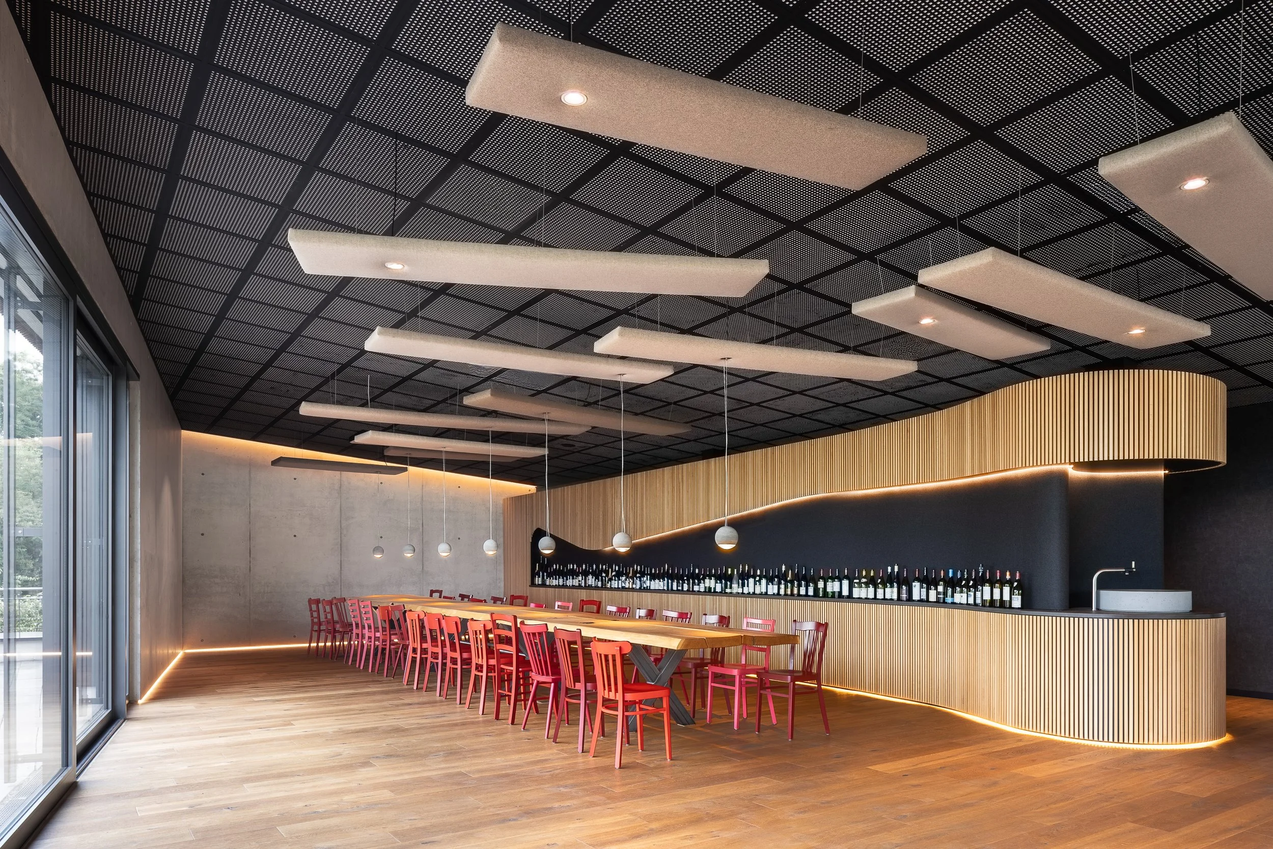 Moderne Innenarchitektur eines Restaurants mit einem hohen, schwarzen, perforierten Deckenpaneel, geschwungenen Holzbar, roten Stühlen und stilvollen Beleuchtungselementen.