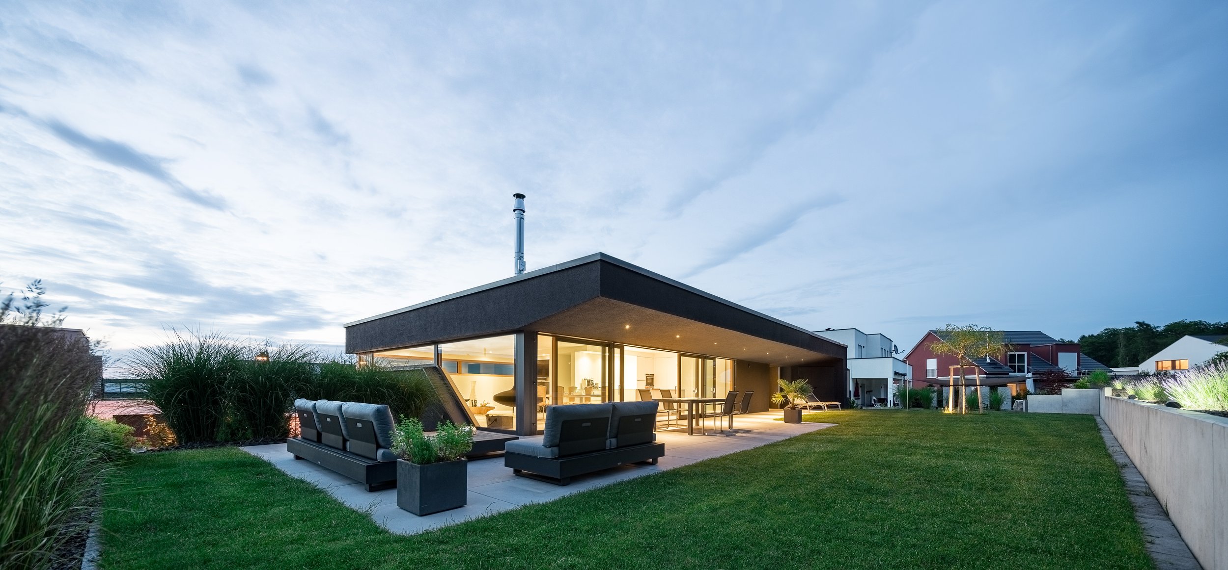 Black Box Haus, Bruchsal, von Henecka Architekten BDA, Moderne Einfamilienhaus mit Terrasse und Garten bei Dämmerung, warmes Licht im Inneren, moderne Architektur, Gartenmöbel, grüne Rasenfläche, hölzerne und betonierte Elemente