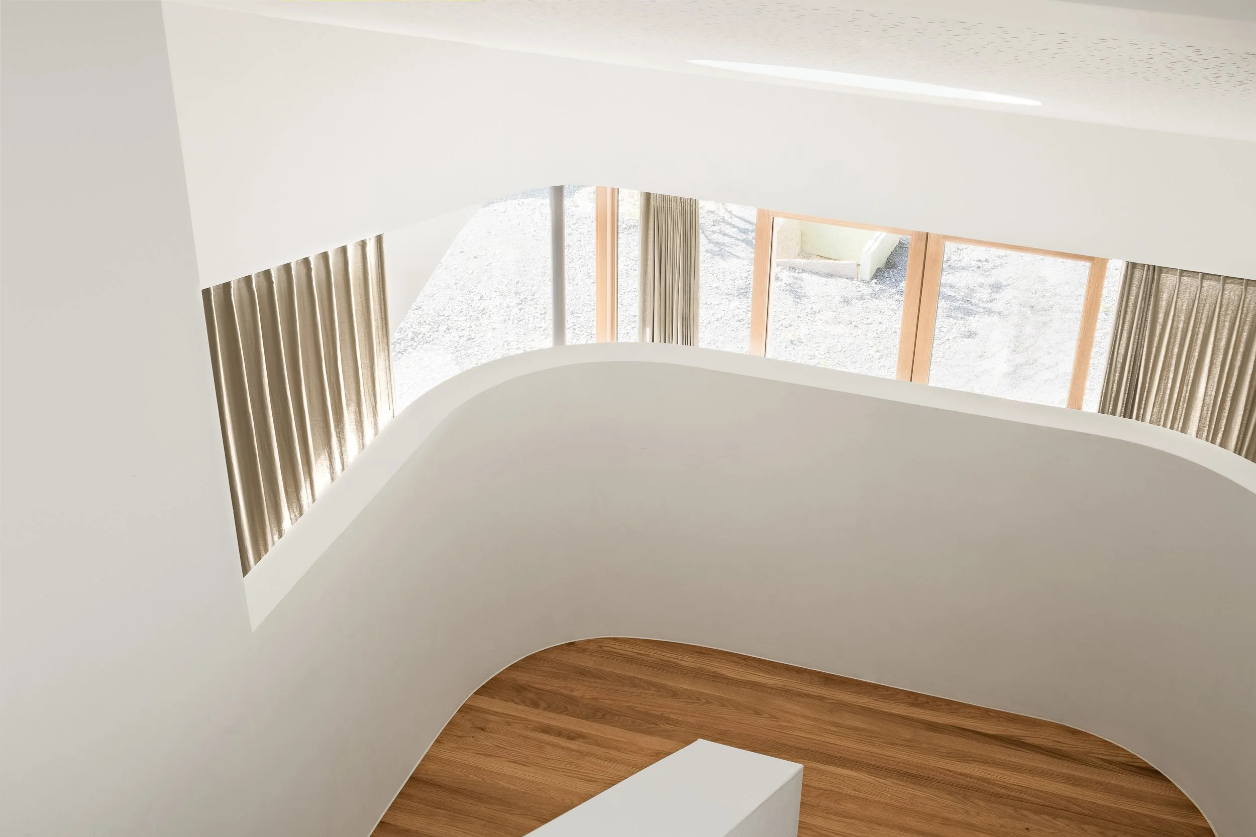 Innenraum mit weißen Wänden, großen Fenstern mit Vorhängen und Holzboden, Blick nach oben auf die Decke.