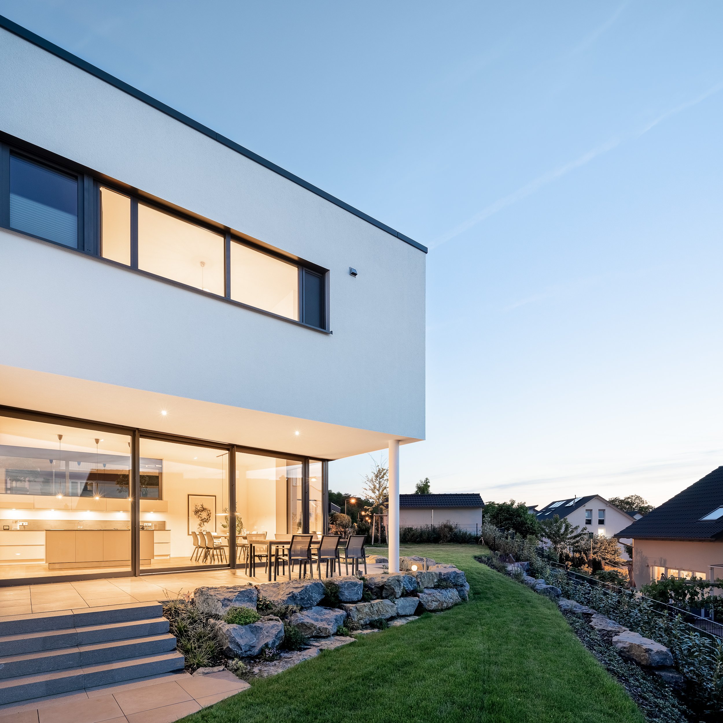 Split Stair Haus Bruchsal von Henecka Architekten BDA Moderne weiße zweistöckige Villa mit großen Fenstern, Terrasse mit Esstisch und Stühlen, umliegender Garten mit Steinen und Rasen, bei Sonnenuntergang
