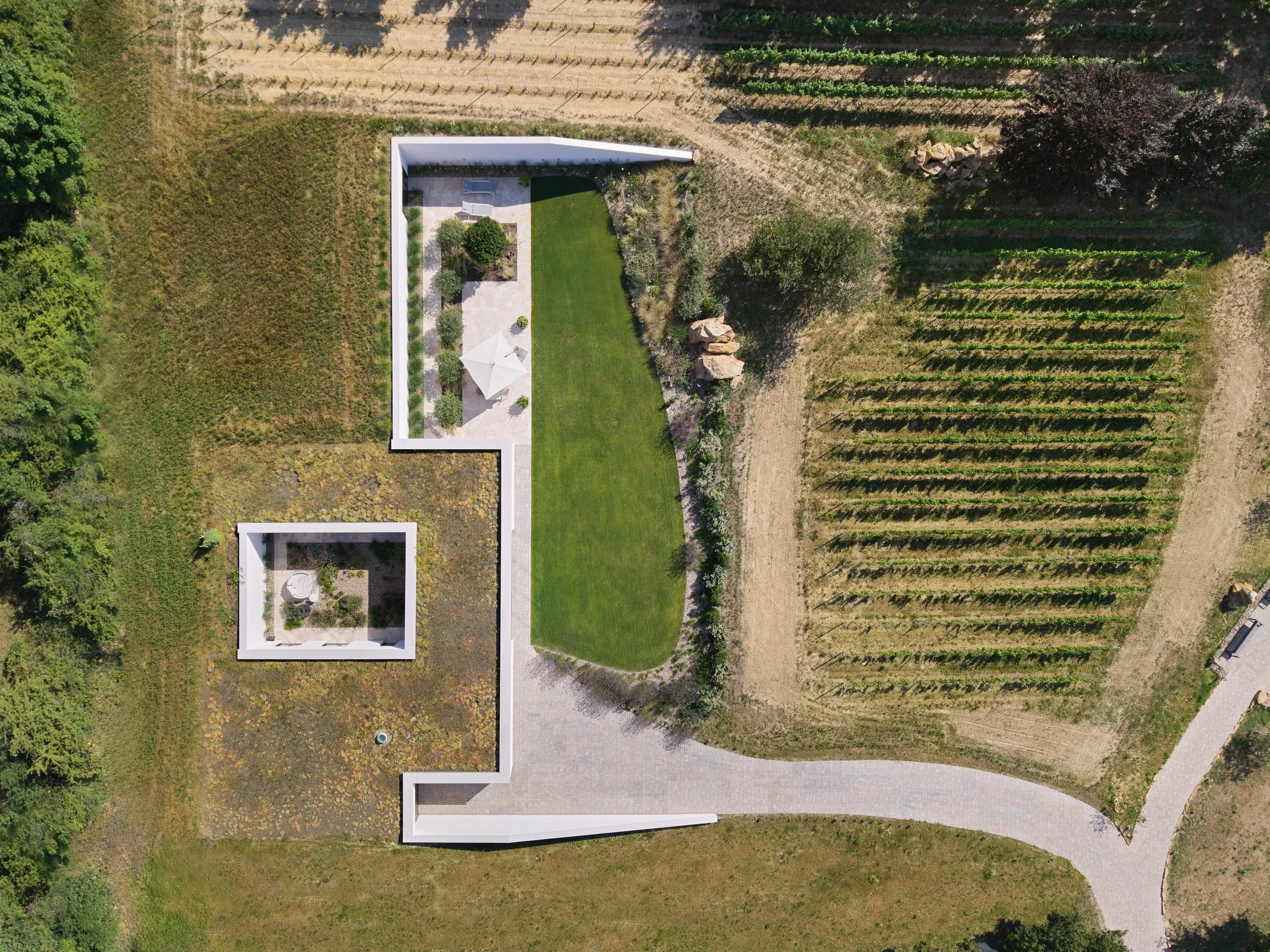 Vogelperspektive auf einen Garten mit grünem Rasen, einer Terrasse mit Sonnenschirm und Stühlen, umgeben von bepflanzten Flächen und angrenzenden landwirtschaftlichen Feldern.