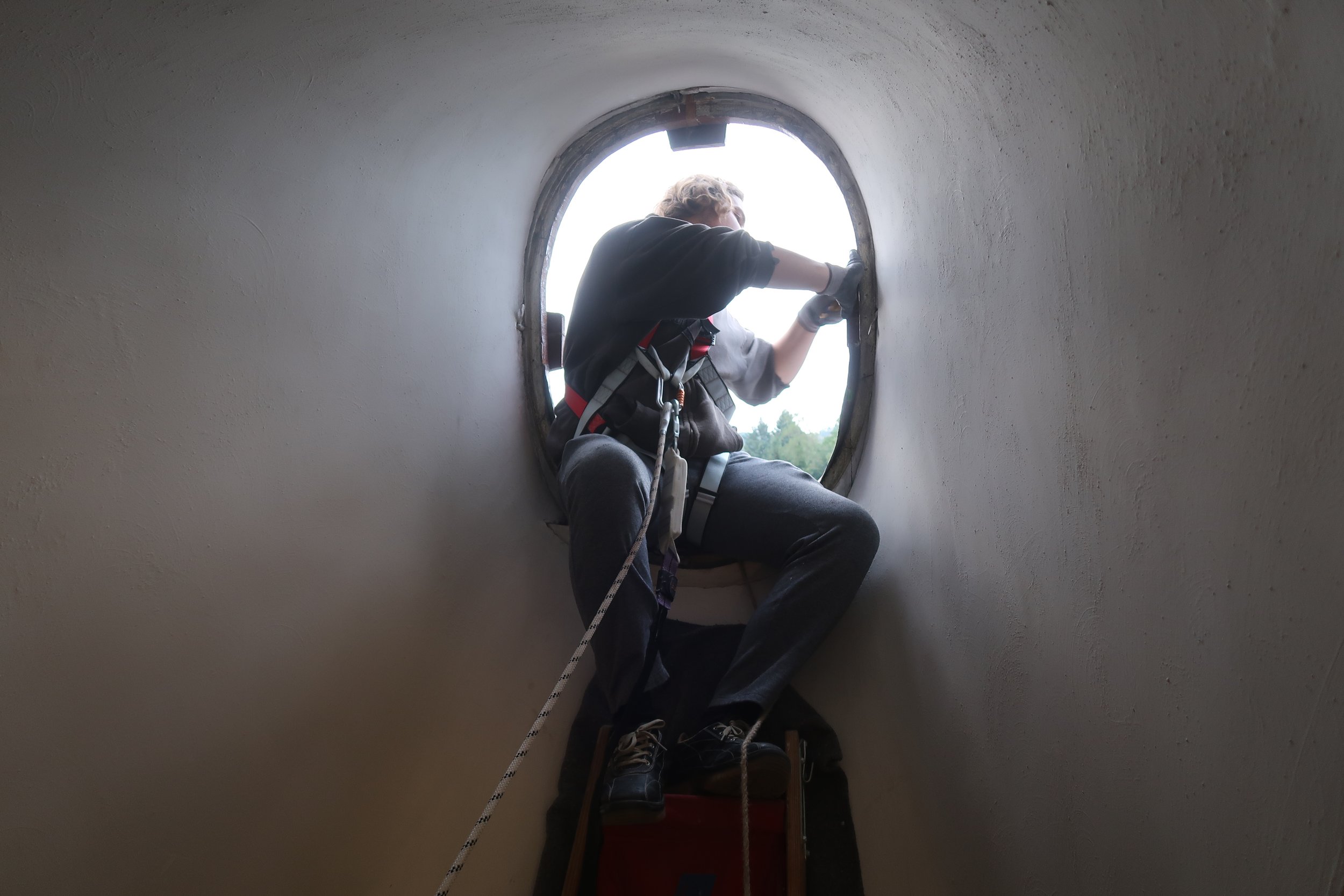 Eine Person klettert durch ein rundes Loch in einer weißen Wand, trägt einen Klettergurt und arbeitet an der Außenwand eines Gebäudes.