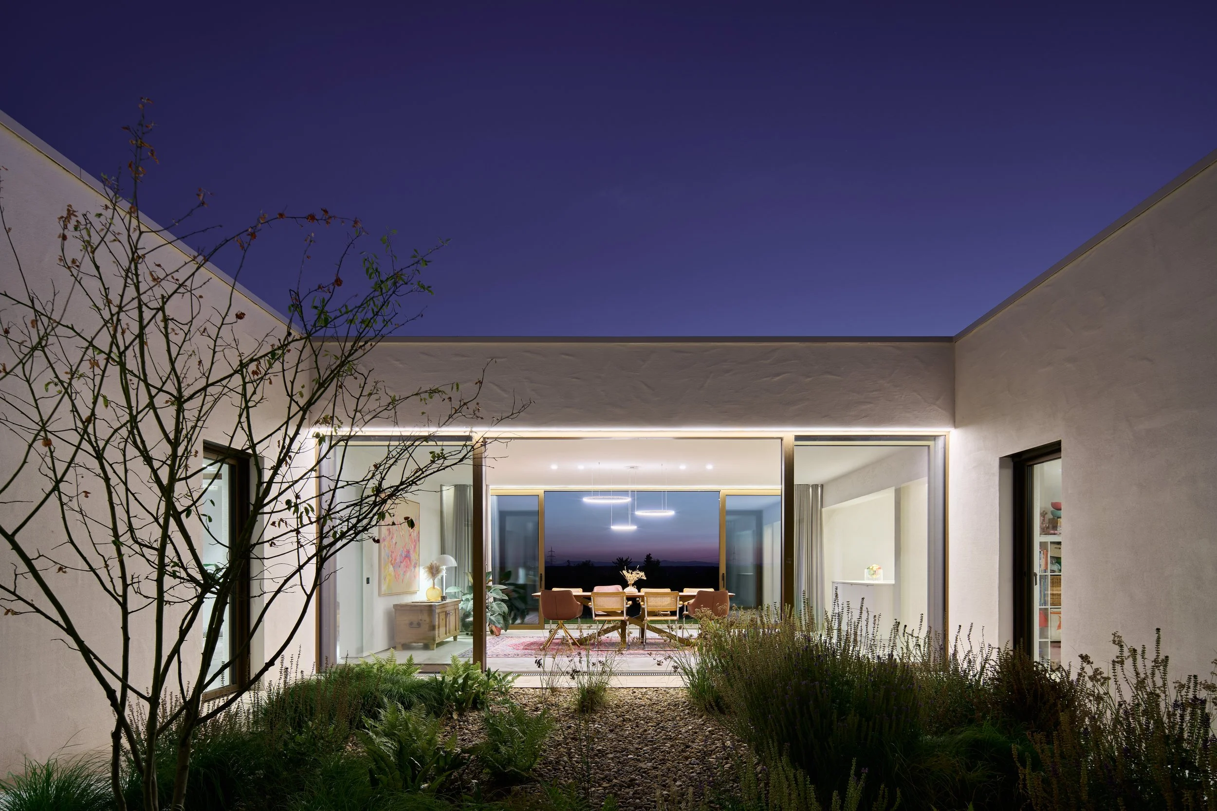 Haus im Weinberg, Bruchsal, von Henecka ArchitekteEin modernes Wohnzimmer mit großen Glasfenstern, Sicht auf den Sonnenuntergang, im Innenraum sind Sessel, Pflanzen und Kunstwerke sichtbar, umgeben von einem Garten mit trockenem Boden und Sträuchern.