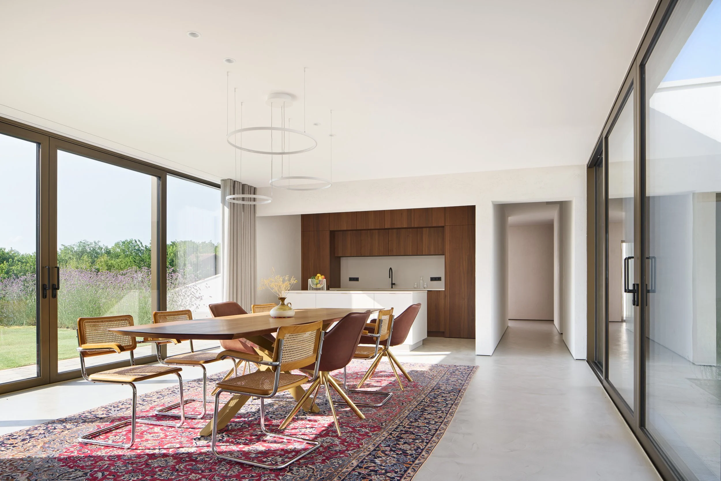 Moderne, offene Wohnküche mit großem Esstisch, Designer-Stühlen, Holzimprägnierung und großen Fenstern, die einen Blick ins Grüne bieten.