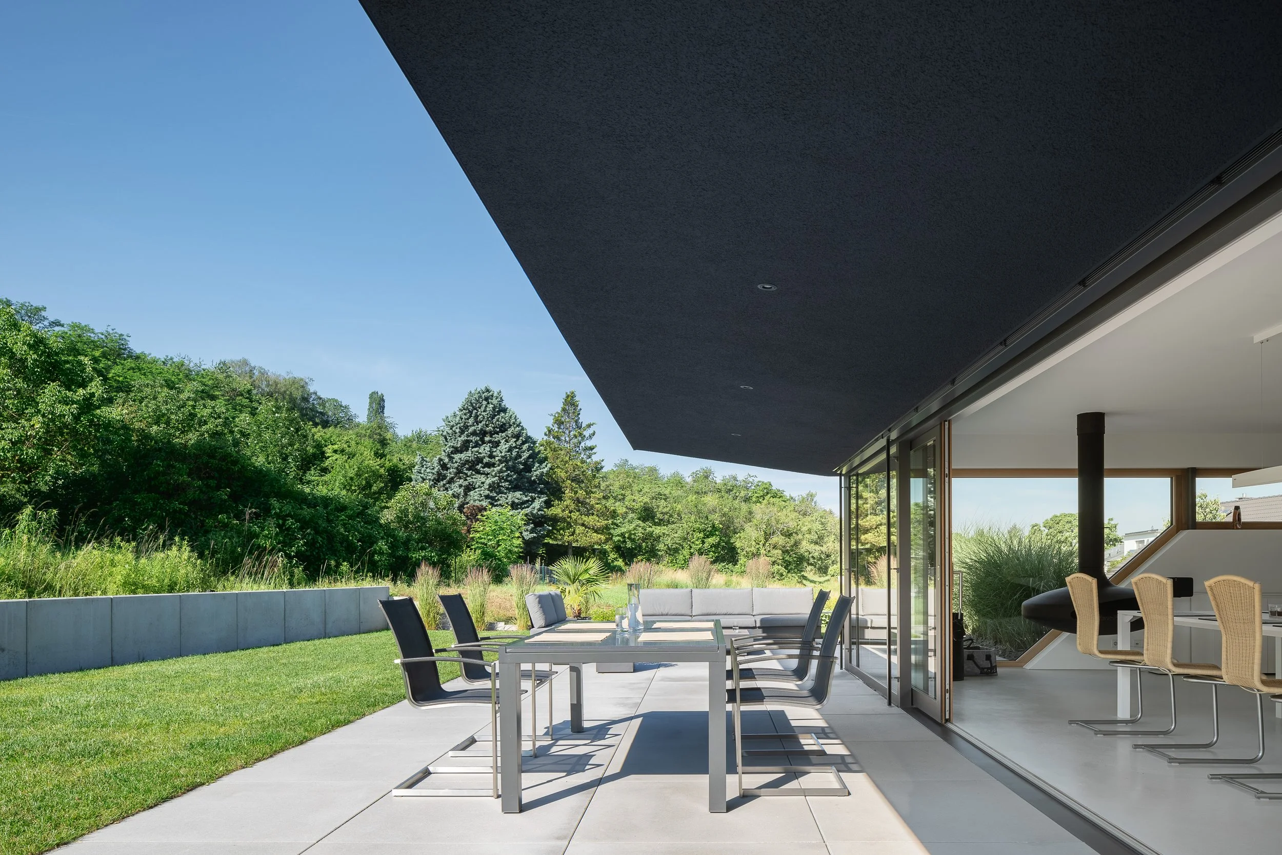 Black Box Haus, Bruchsal, von Henecka Architekten BDA, Moderner Außenbereich eines Hauses mit Terrassenmöbeln, großen Glasfenstern und Blick auf einen grünen Garten mit Bäumen bei sonnigem Himmel.