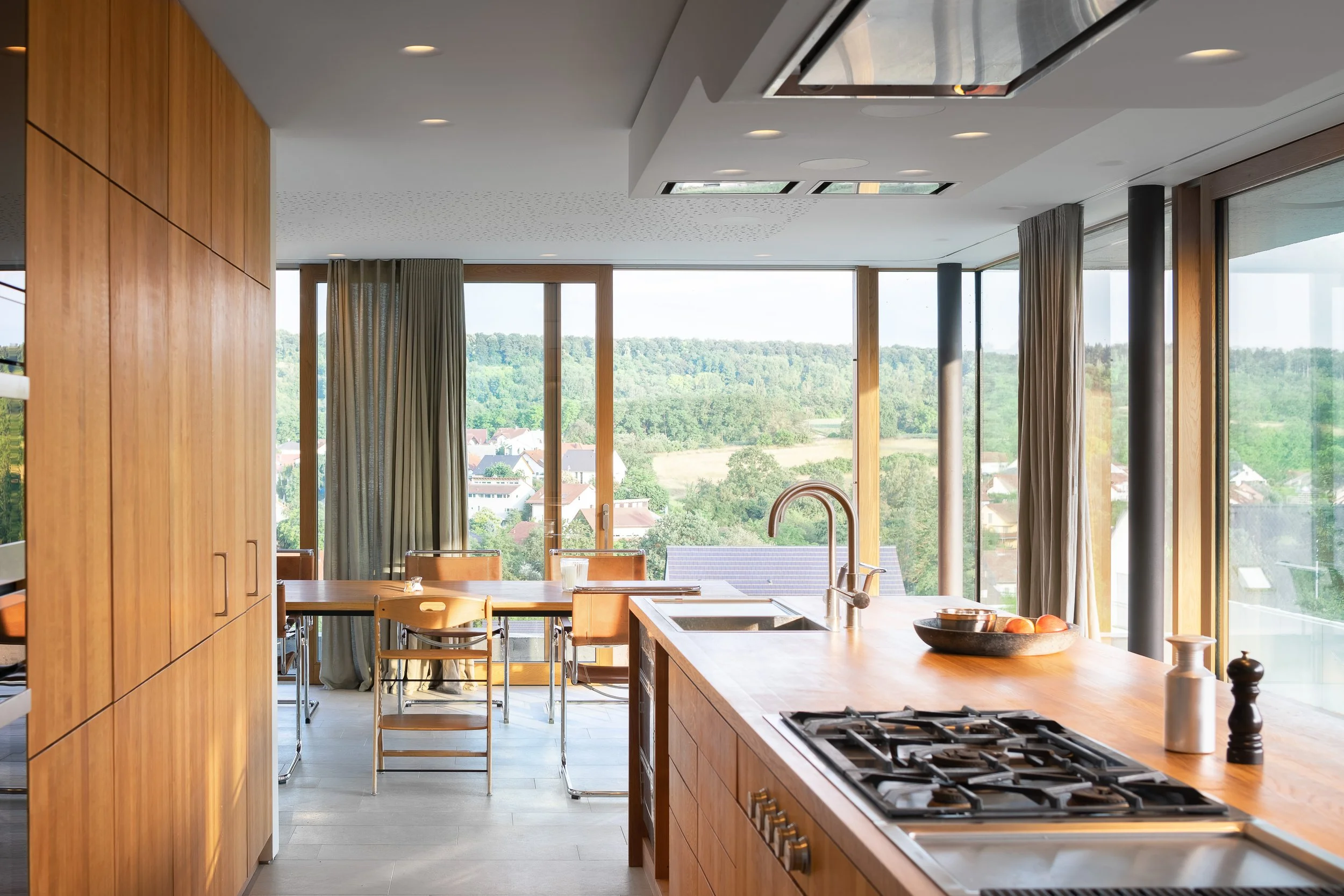 Moderne, helle Küche mit Holzmöbeln, großer Fensterfront mit Blick auf eine grüne Landschaft, Esstisch mit Stühlen, Kücheninsel mit Herd, Lavabro und Obst auf der Insel.