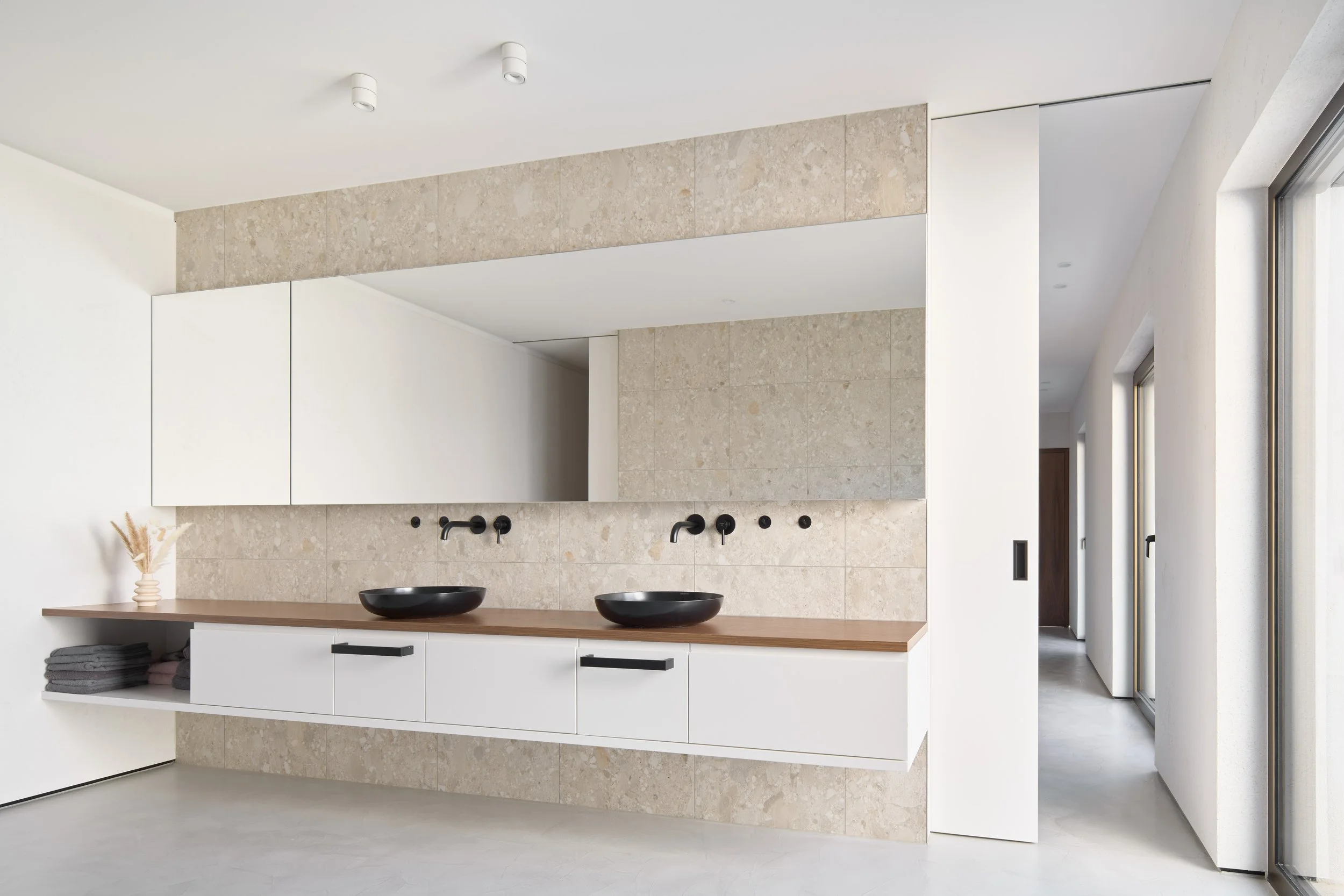 Moderner minimalistischer Waschraum mit zwei schwarzen Waschbecken, weißen Schränken, beige Fliesen und großen Fenstern, die viel natürliches Licht hereinlassen.