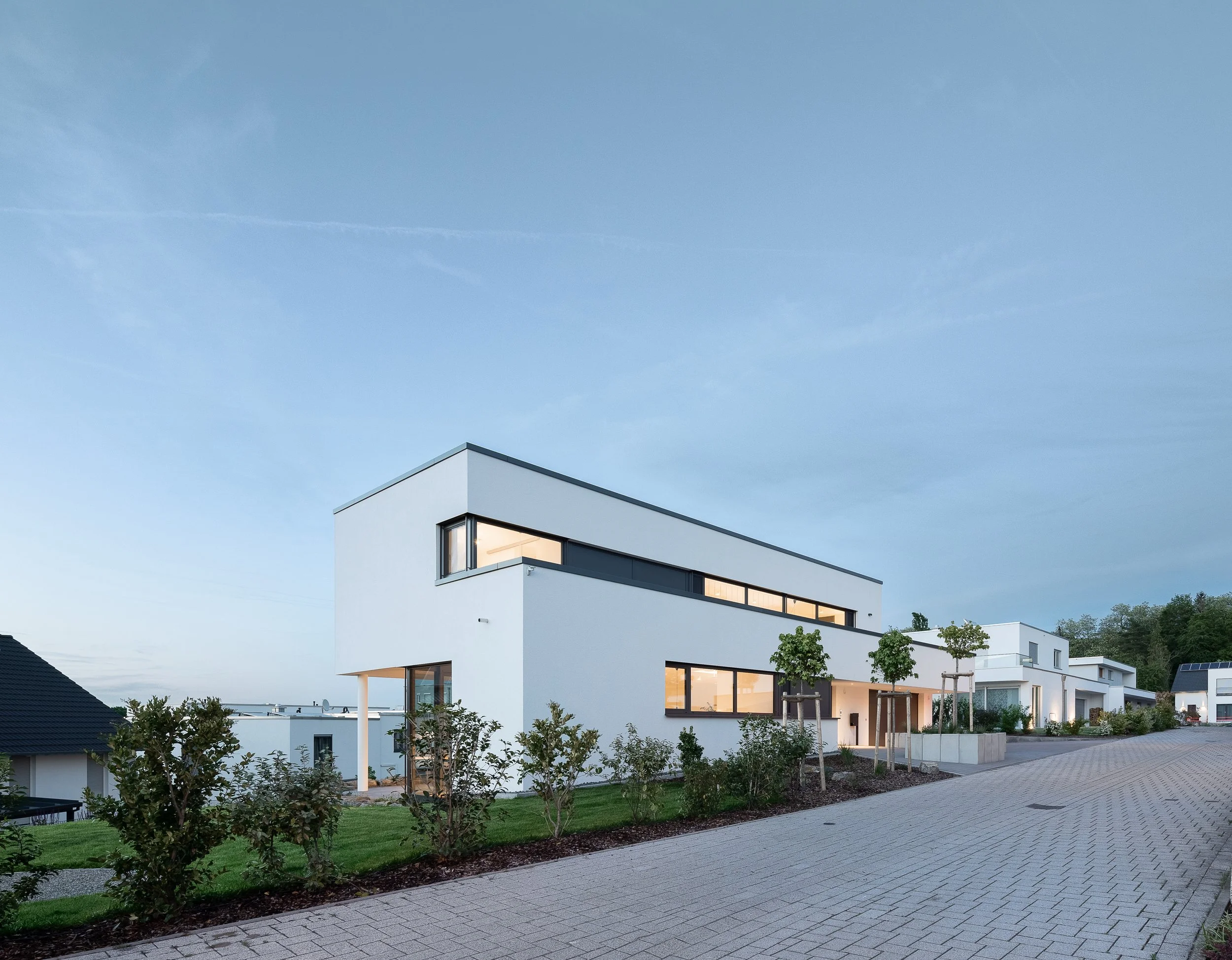 Modernes, weißes Mehrfamilienhaus mit großen Fenstern und Balkon, umgeben von grünem Garten und Gehweg, bei Sonnenuntergang.
