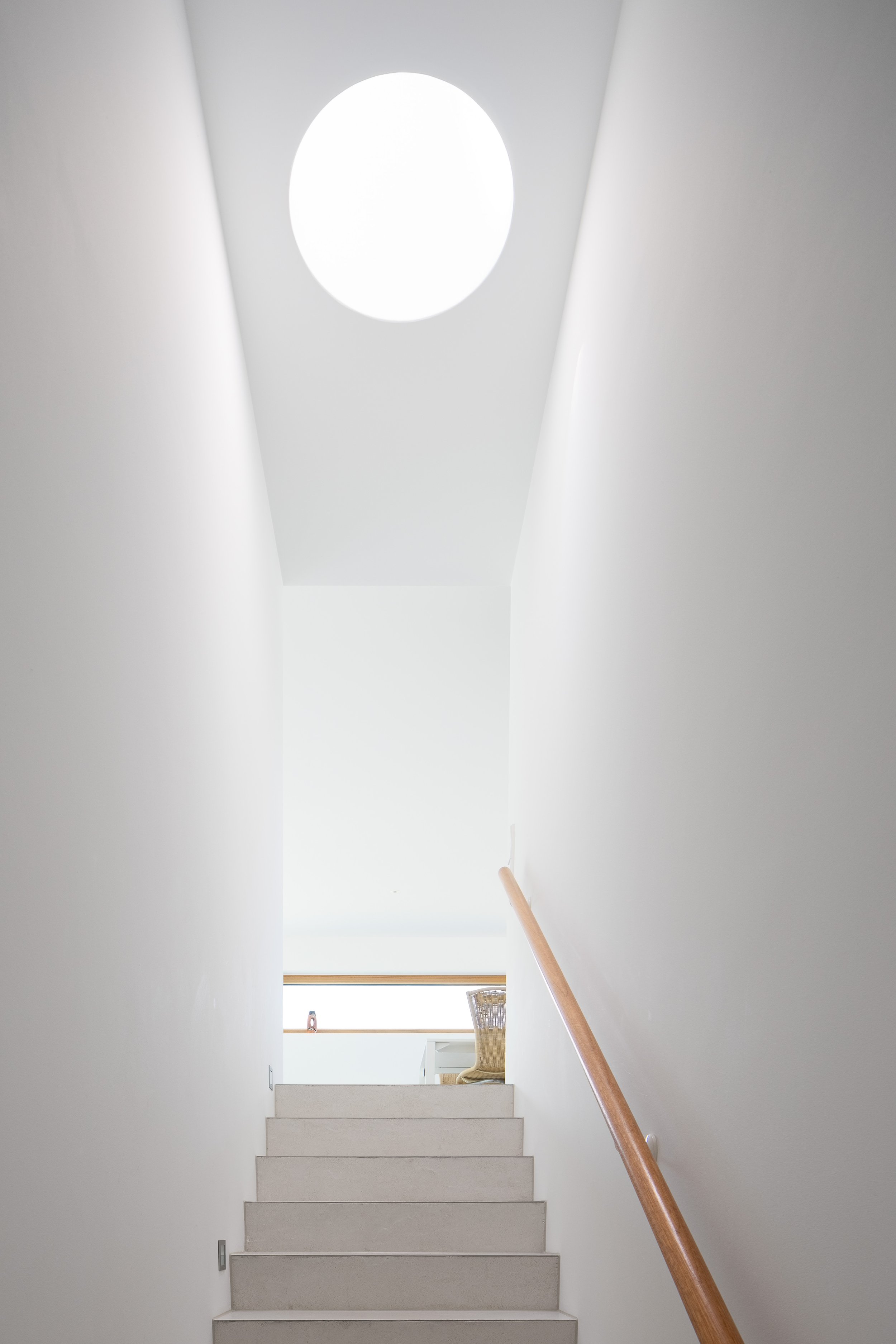 Black Box Haus, Bruchsal, von Henecka Architekten BDA, Innenaufnahme eines hellen Treppenhauses mit weißen Wänden, weißen Stufen, Holzhandlauf und einem Balkon am oberen Ende mit einem Stuhl und einer Lampe.
