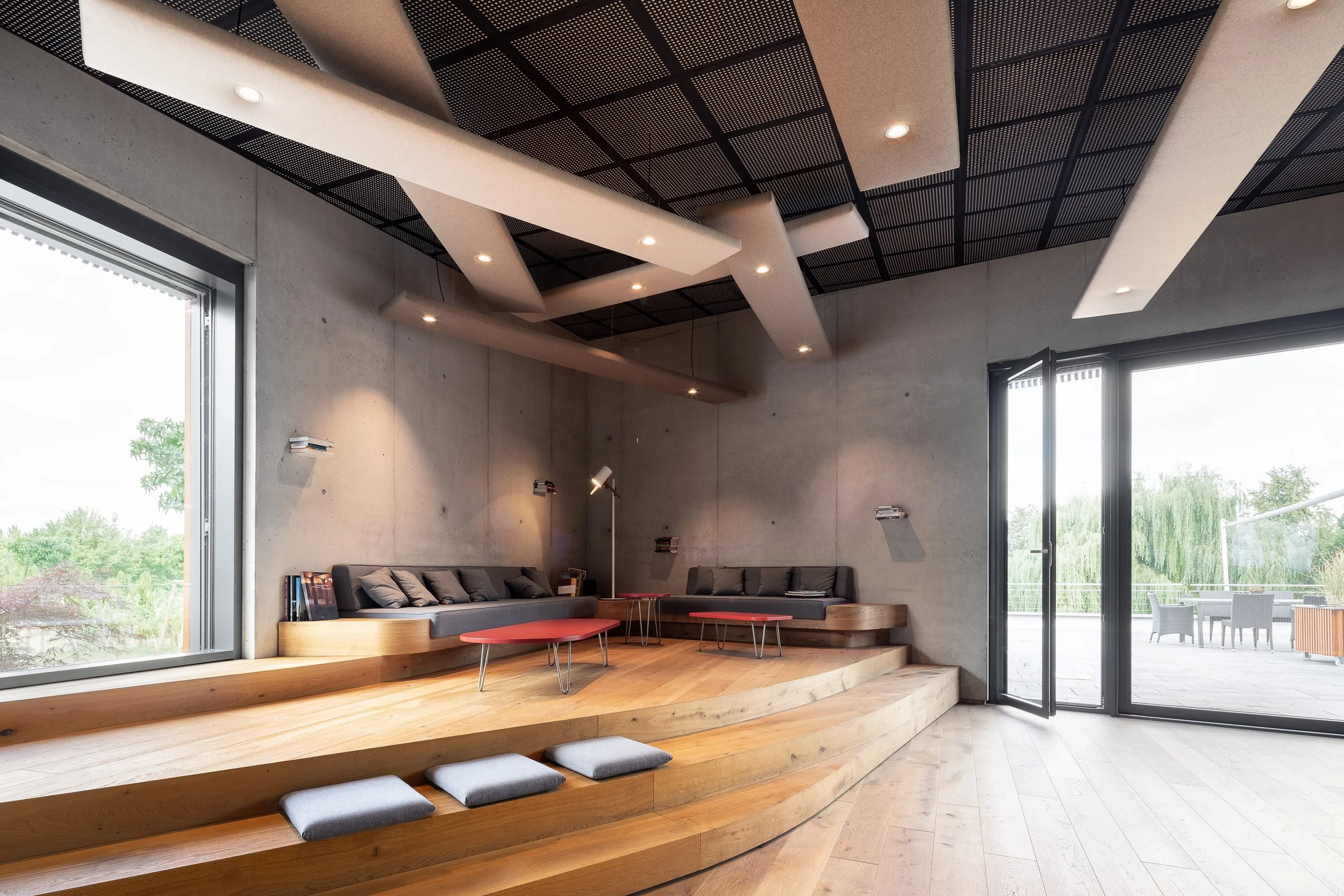 Innenraum eines modernen Raumes mit großen Fenstern, Holzböden, grauem Sofa, roten Tischen und einer schwarzen Deckenverkleidung mit Einbaulampen.