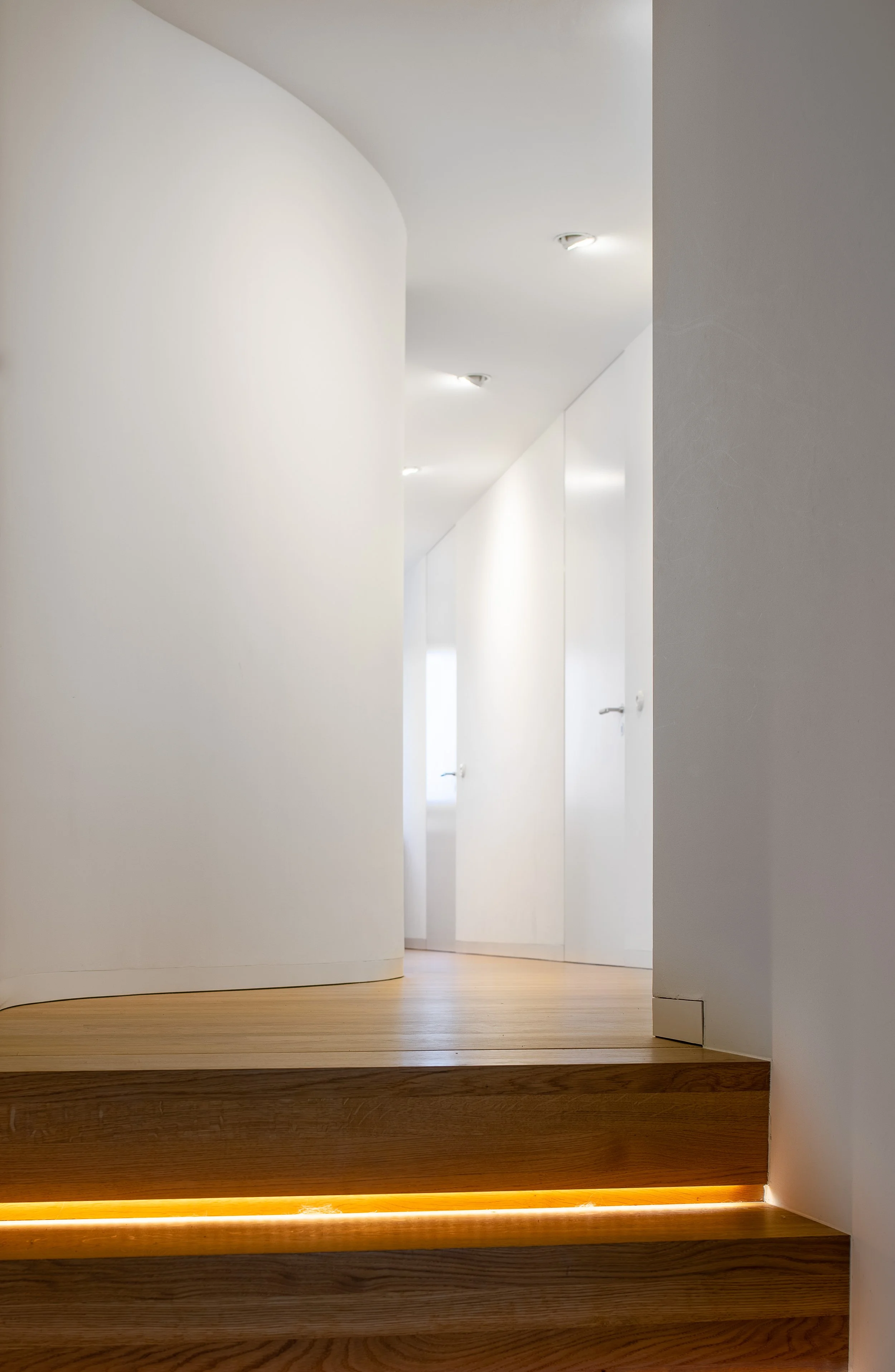 Innenaufnahme eines weißen Flurs mit gewölbter Decke und Holztreppe, beleuchtet durch eingebautes Licht.