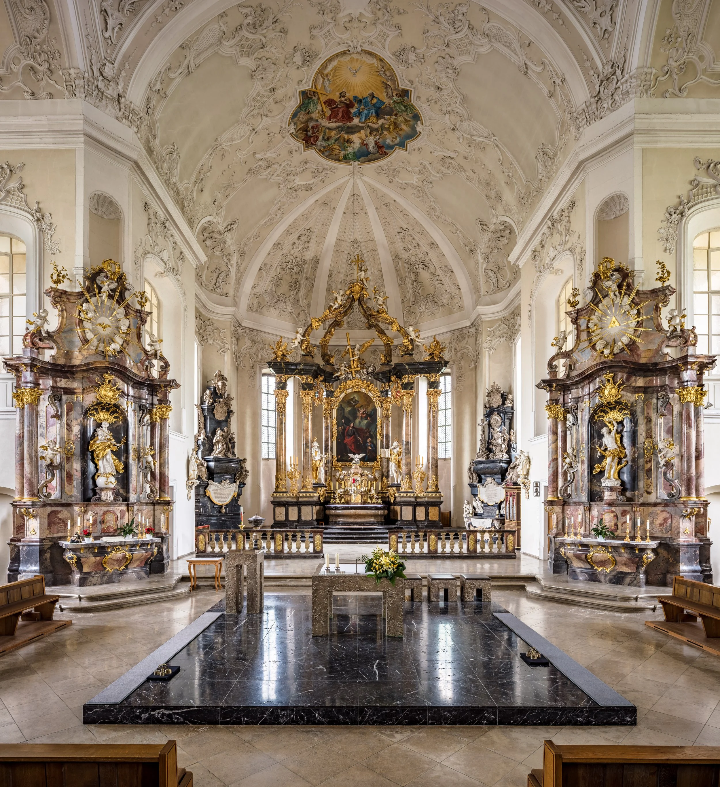 Innenraum einer barocken Kirche mit geschnitztem Altar, barocken Statuen, goldverzierten Details und einem Deckenfresko, das den Himmel mit Engeln zeigt.