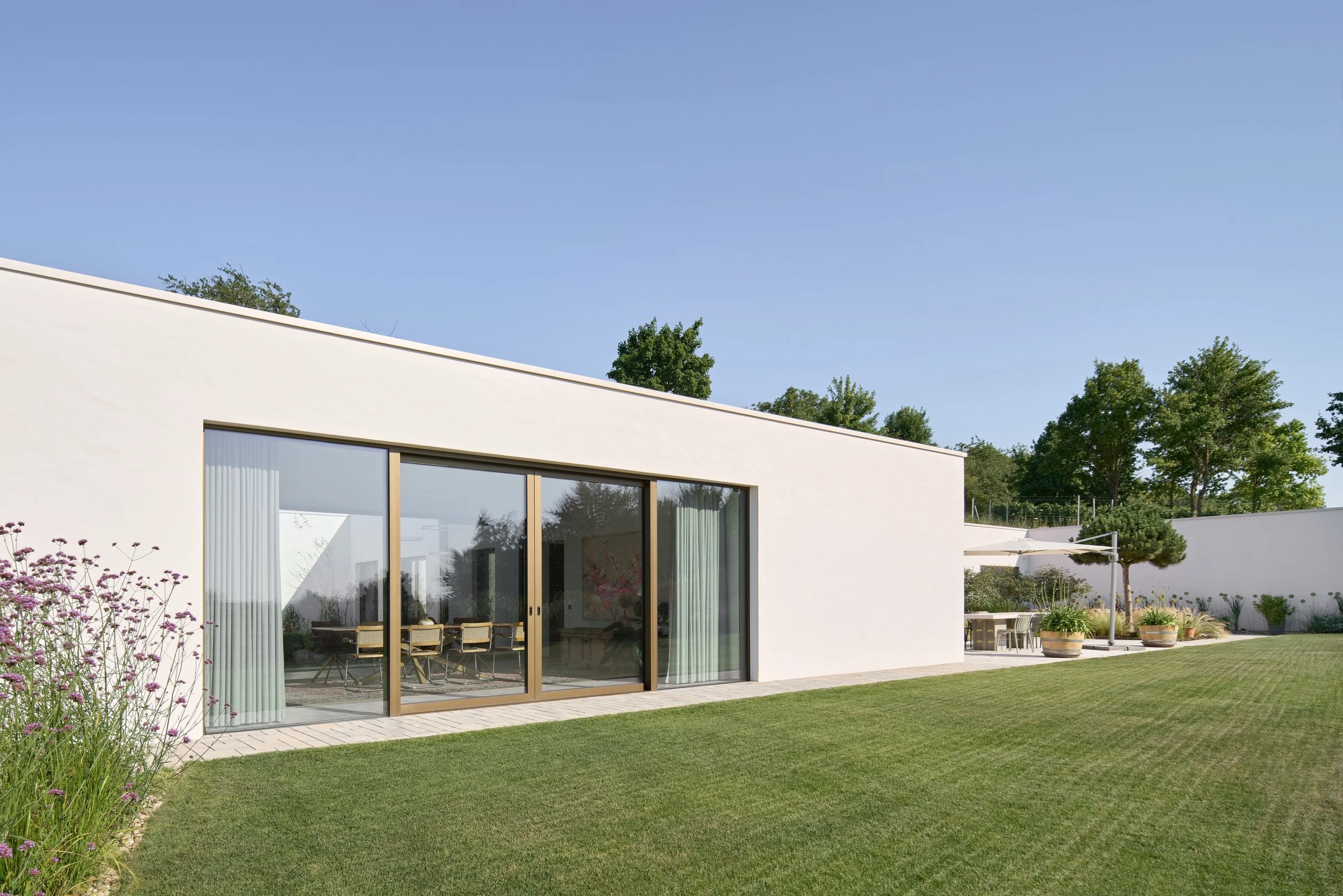 Haus im Weinberg, Bruchsal, von Henecka Architekten BDA, Moderne weiße Hausfassade mit großen Glasfenstern, umgeben von einem Garten mit grünem Rasen, Sträuchern und Bäumen, bei sonnigem Himmel.