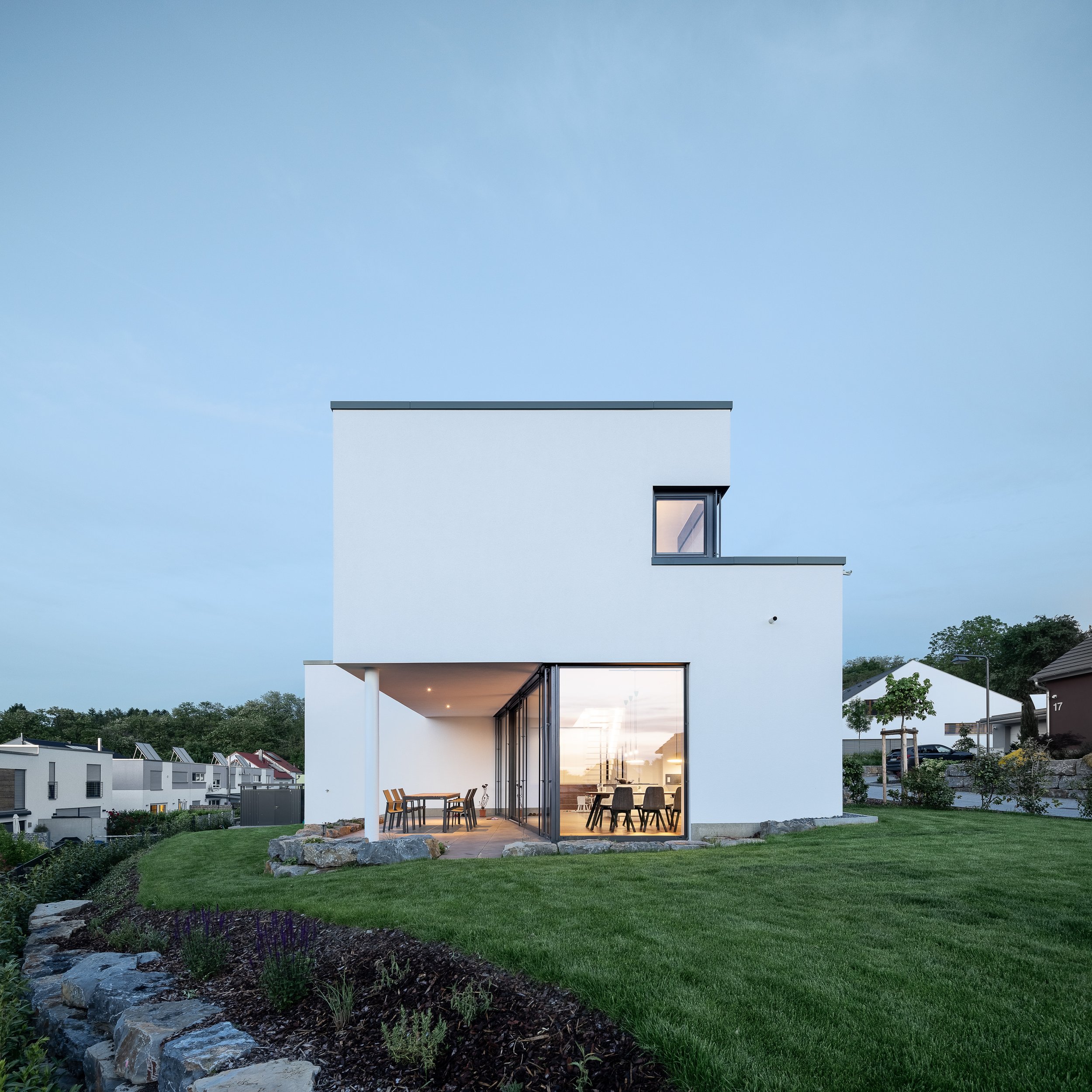 Split Stair Haus, Bruchsal, von Henecka Architekten BDA, Modernes Haus mit großen Fenstern und Terrasse, umgeben von Garten mit Rasen, Blumen und Bäumen, bei Sonnenuntergang.
