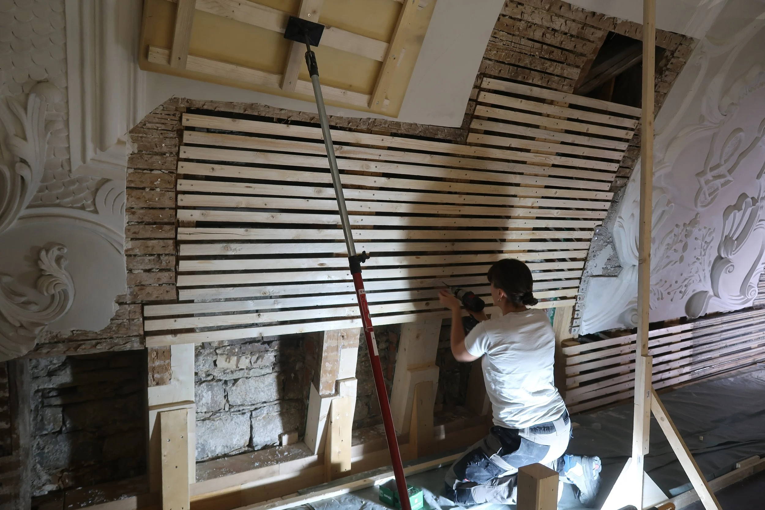 Eine Frau arbeitet an einem Baugerüst innerhalb eines Gebäudes mit verzierten Wanddekorationen. Sie benutzt ein Elektrowerkzeug und ist in handwerklicher Arbeit beschäftigt.