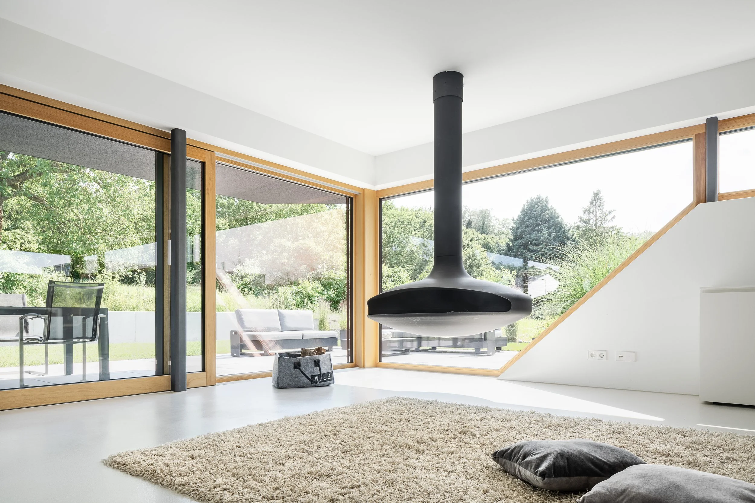 Black Box Haus, Bruchsal, von Henecka Architekten BDA, Moderner Wohnraum mit großen Fenstern, einem schwarzen Kamin, Teppich und Kissen, Blick ins Grüne nach draußen.