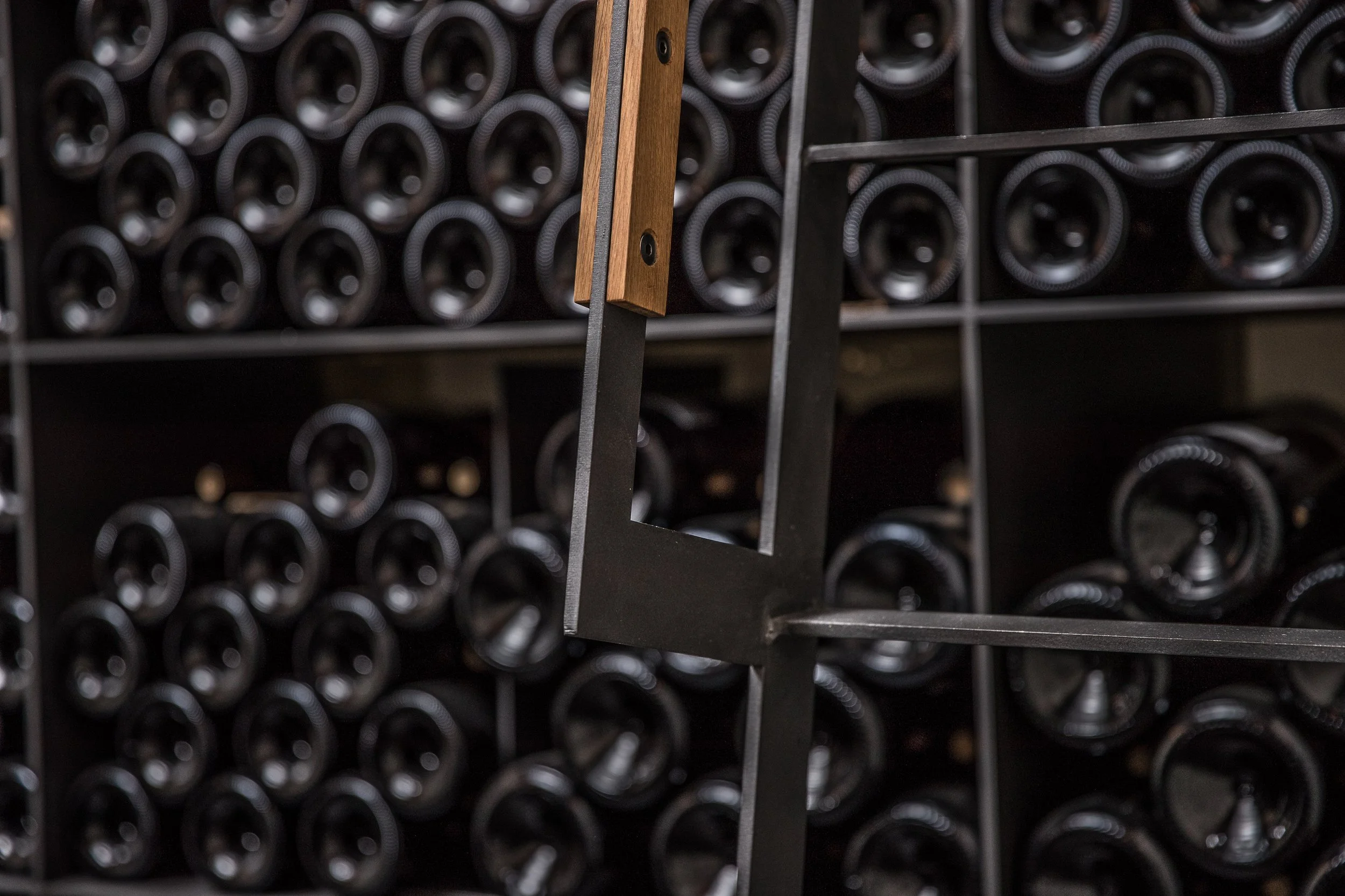 Vinothek Weingut Klumpp Bruchsal von Henecka Architekten BDA, Blick auf eine Weinflaschenablage mit mehreren schwarzen Flaschen, teilweise durch einen schwarzen Metallrahmen sichtbar, und einem Holzstück als Dekorations- oder Befestigungselement.