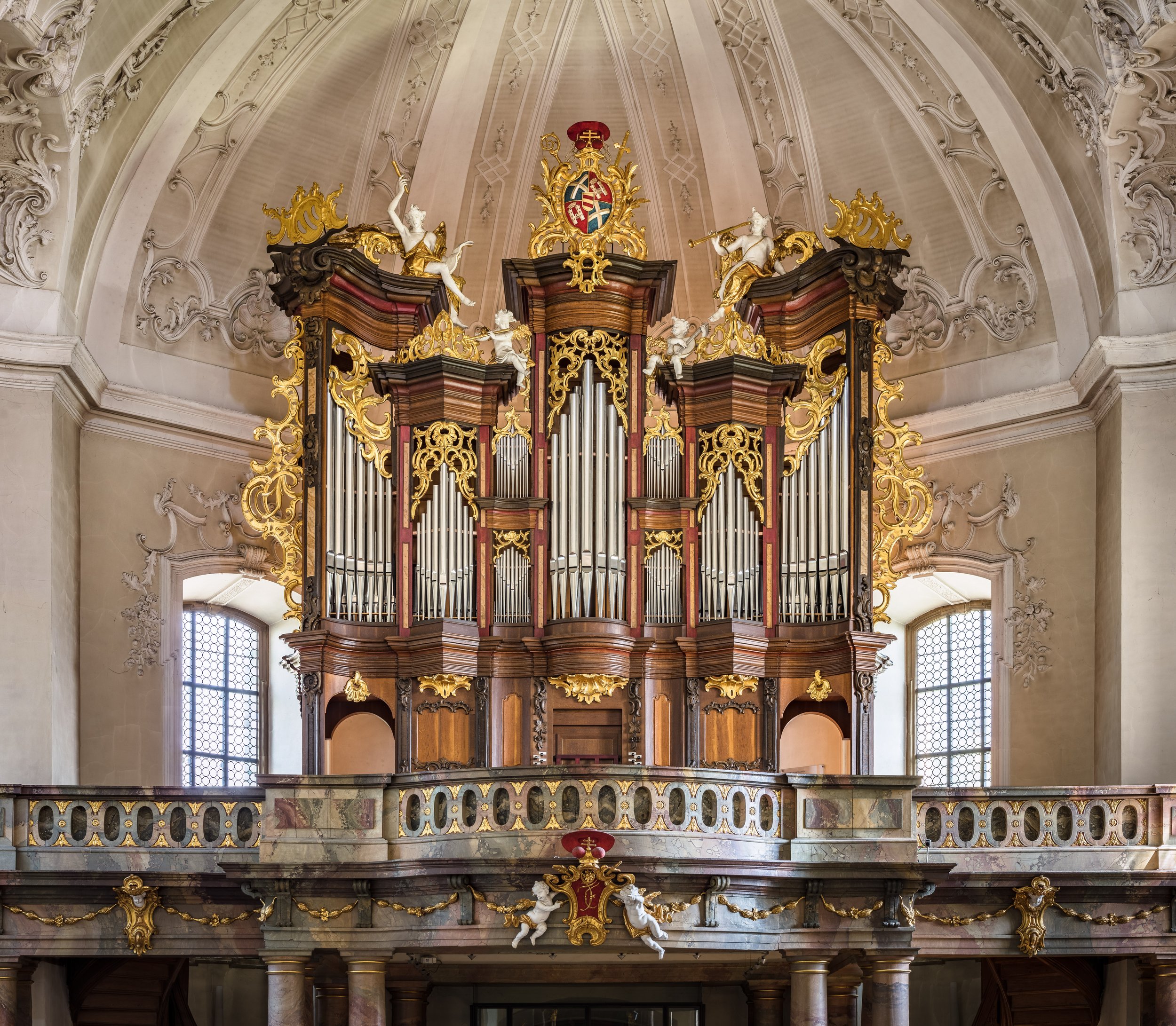Barock-Orgel mit aufwändiger goldener Verzierung, Skulpturen und einem Wappen unter einer hohen, formelhaften gewölbten Decke in einer Kirche, mit Fenstern im Hintergrund.