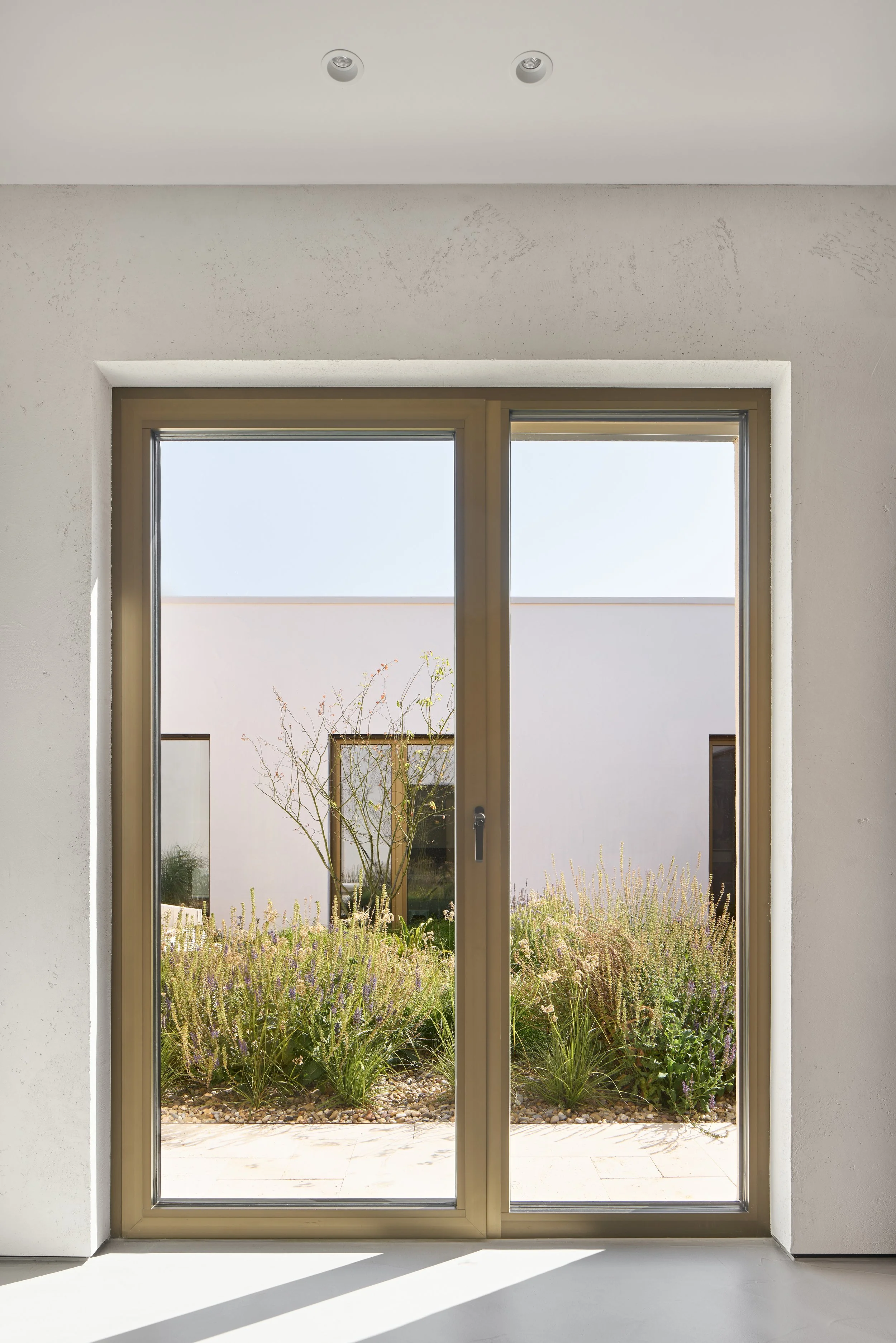Blick durch eine große Glastür auf einen Garten mit Blumen und einem Baum.