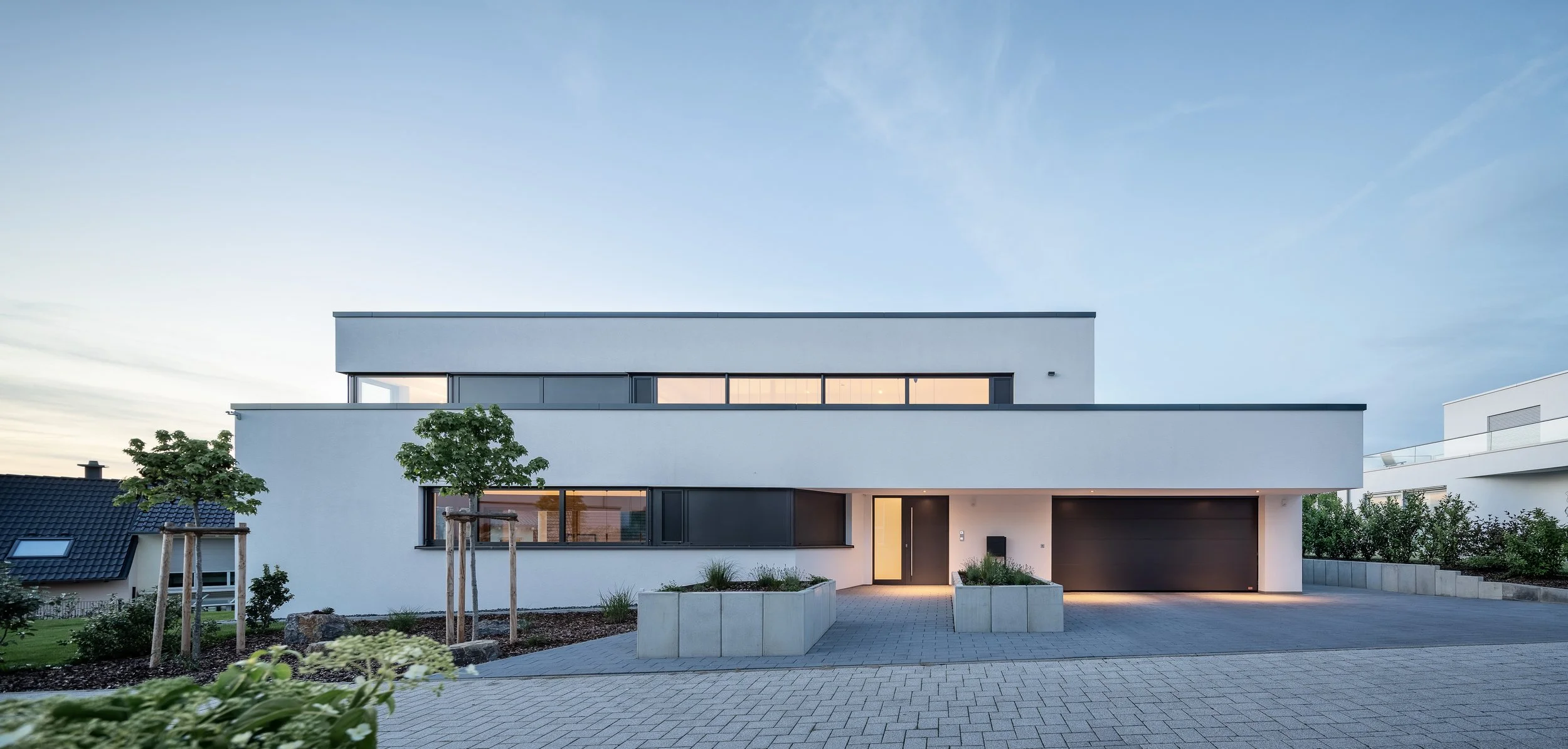 Split Stair Haus Bruchsal von Henecka Architekten BDA Modernes weißes Haus mit mehreren Etagen, großen Fenstern und Garage, umgeben von einer gepflasterten Einfahrt, Bäumen und Garten, bei Dämmerung.