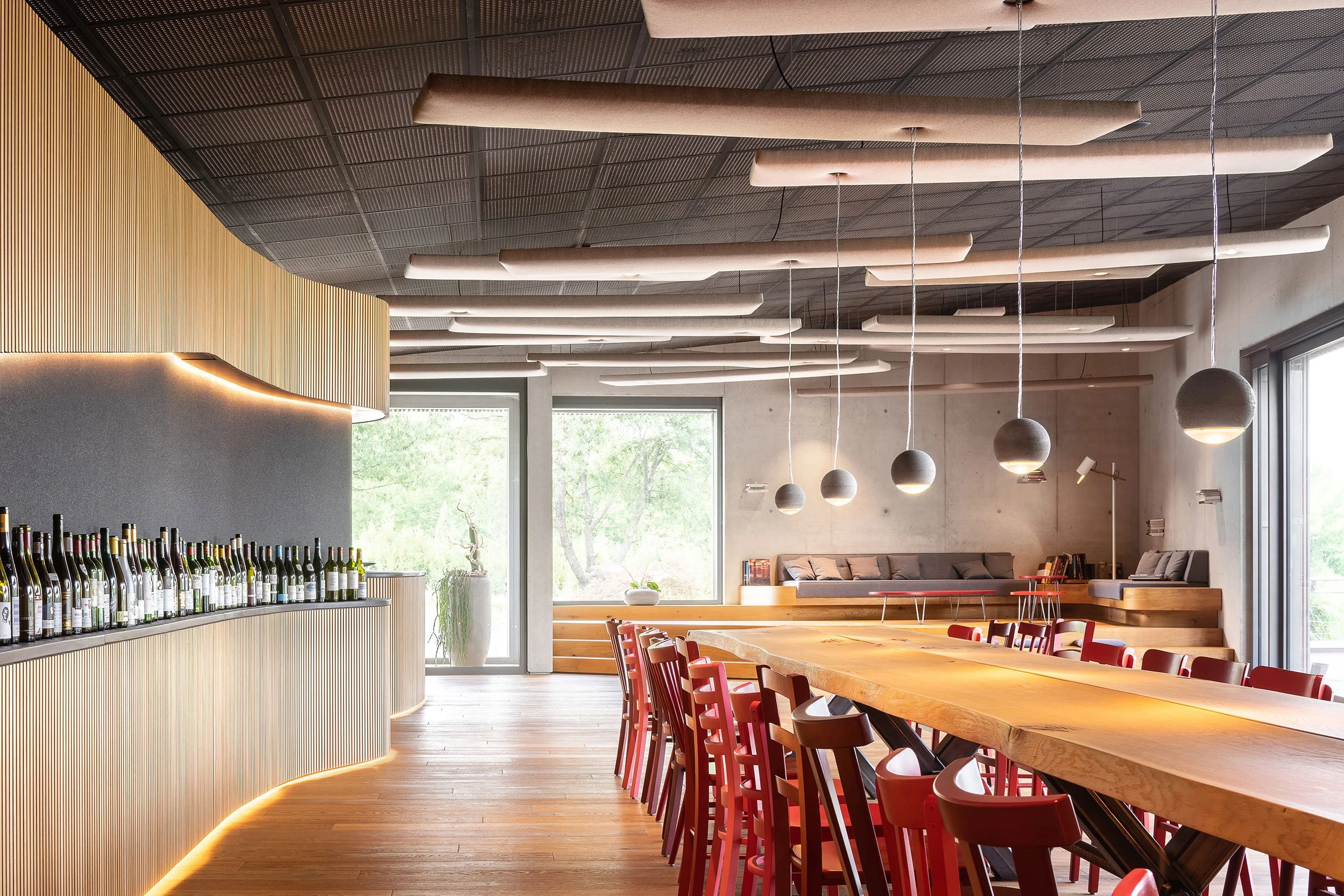 Innenaufnahme eines modernen, stilvollen Restaurants mit einem langen Holztisch, roten Stühlen und großen Fenstern, die natürliches Licht hereinlassen.