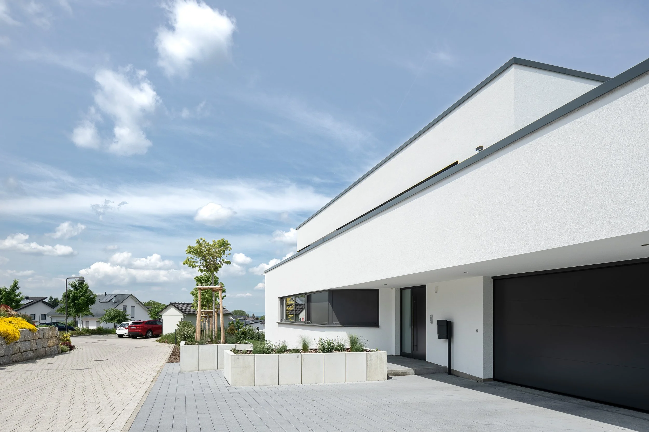 Modernes, weißes Wohnhaus mit Garage, vor dem Haus befinden sich ein gepflasterter Weg und eine offene Terrasse. Im Hintergrund sind weitere Häuser und Bäume bei sonnigem Himmel zu sehen.