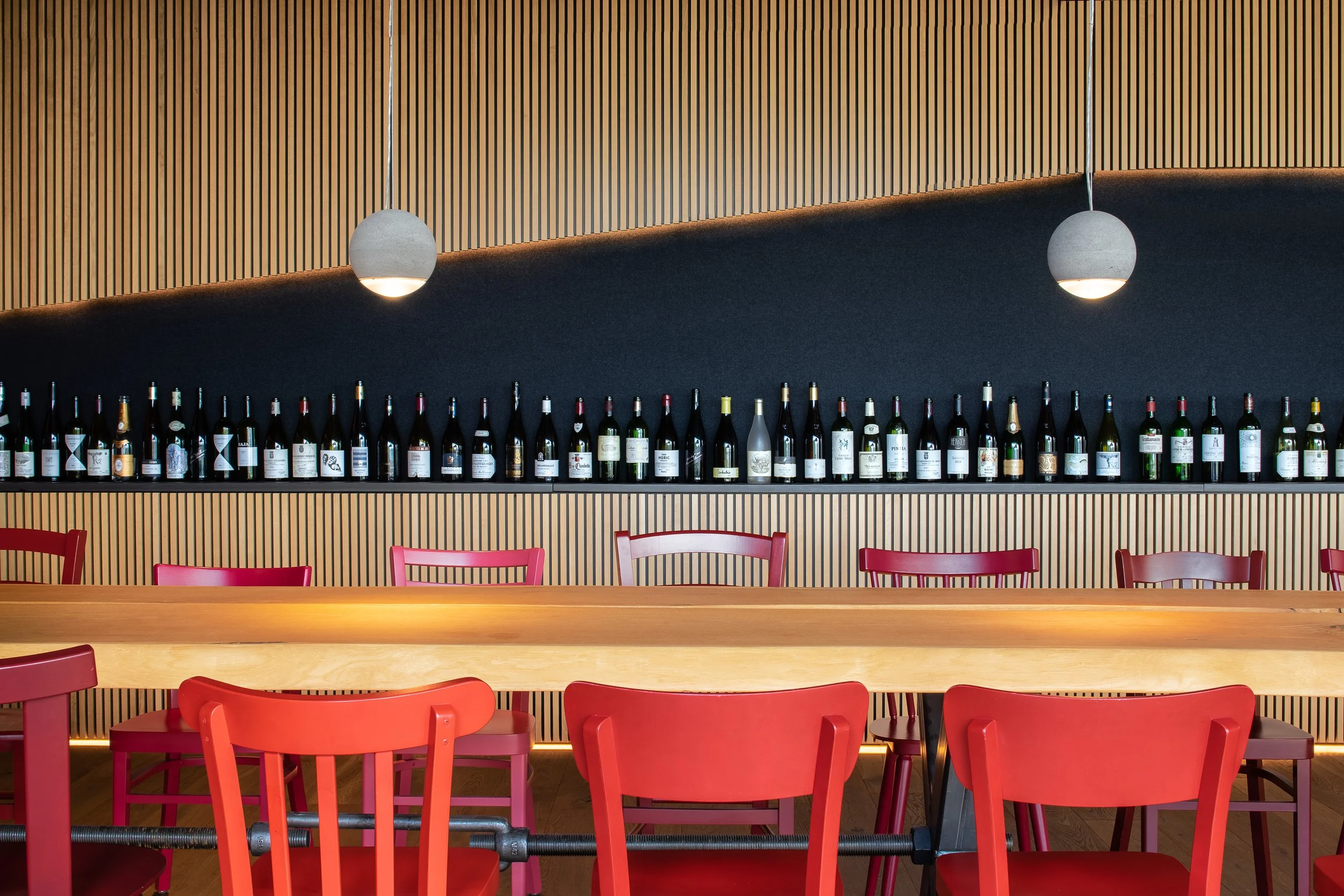 Innenraum eines Restaurants mit einer langen Regal mit verschiedenen Weinflaschen, roten Stühlen und zwei weißen Pendelleuchten an einer Holzpaneelwand.