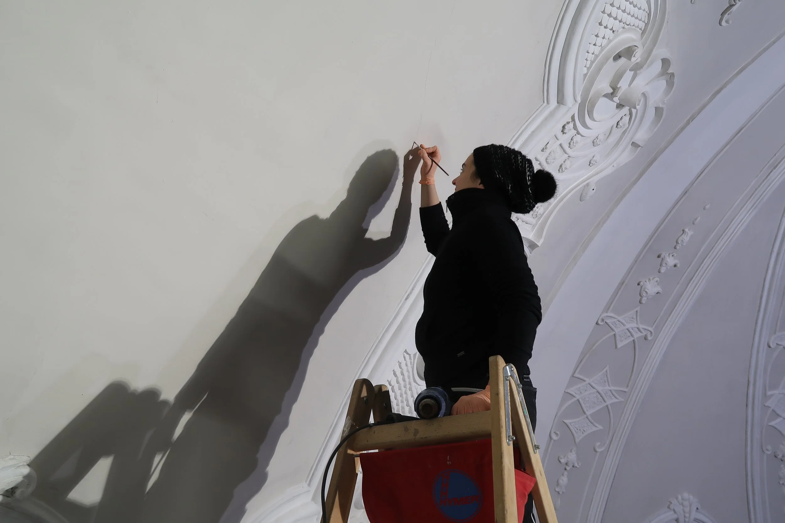 Eine Frau steht auf einer Leiter und malt an einer weißen Wand in einem Raum mit dekorativen Wandverzierungen, vermutlich im Rahmen von Renovierungsarbeiten.