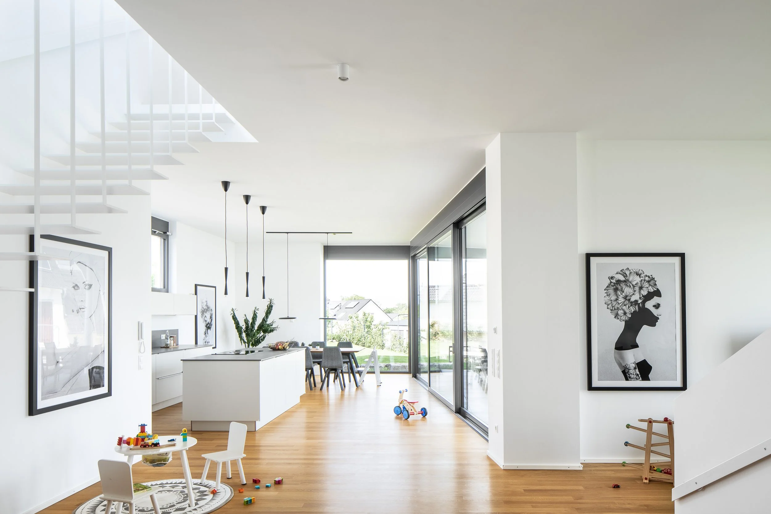 Split Stair Haus, Bruchsal, von Henecka Architekten BDA, Ein modern eingerichteter, heller Wohnraum mit offener Küche, Esstisch, Spielzeug für Kinder und Kunstwerken an den Wänden.
