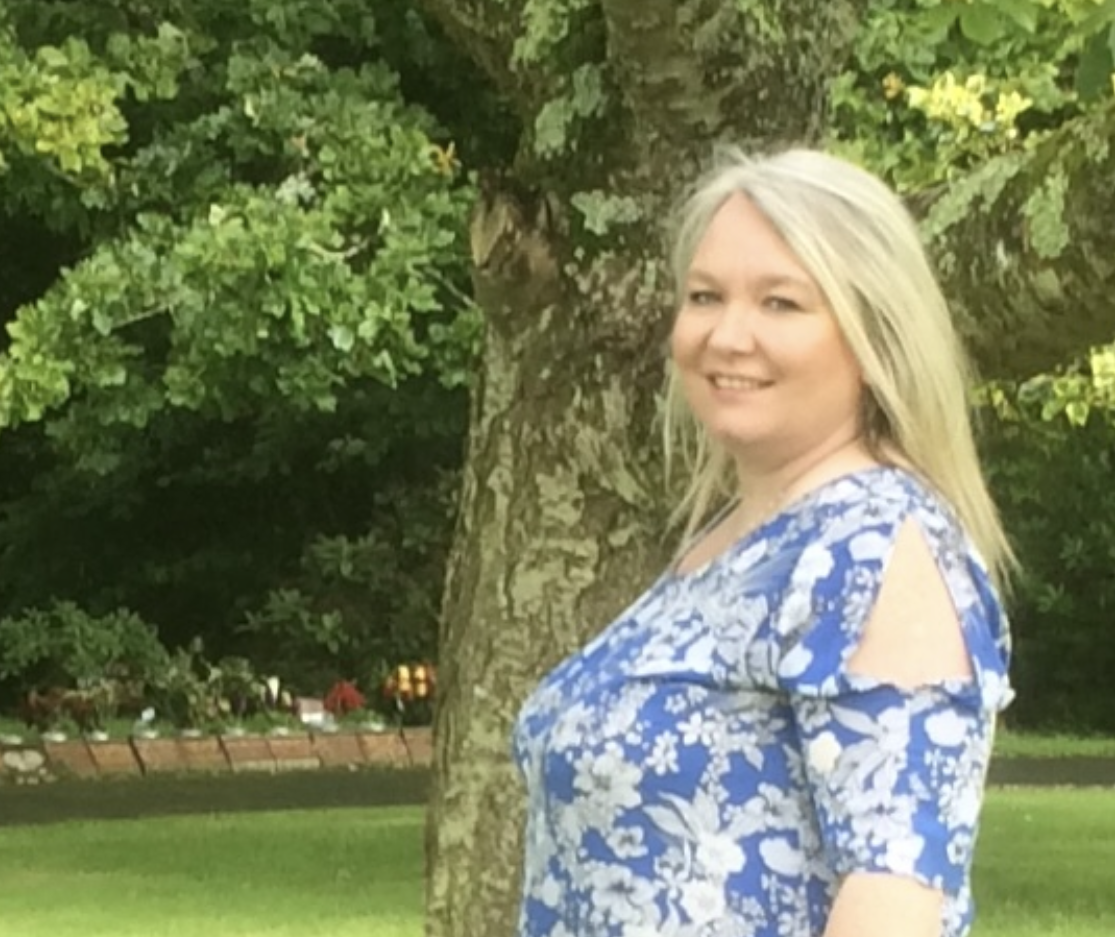 A smiling woman with long blonde hair wearing a blue floral dress stands outdoors near a tree, with greenery and a garden area in the background.