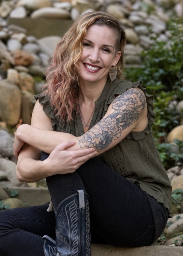 A woman with wavy, pinkish-blonde hair, smiling and sitting outdoors among rocks and greenery, wearing a sleeveless dark green top, a tattooed arm, black pants, and black boots.