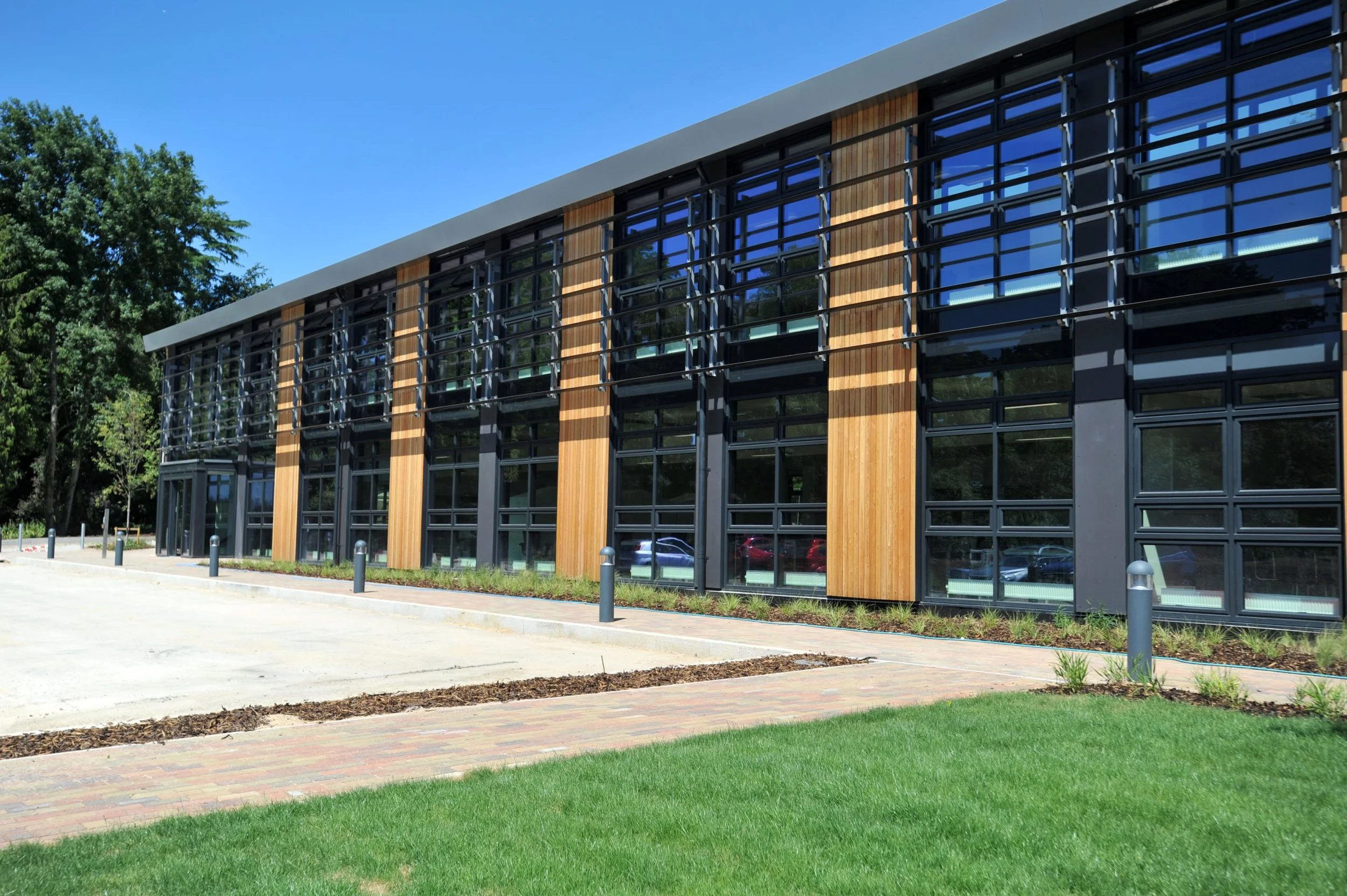 Modern two-story building with glass windows, wooden accents, and outdoor lighting, surrounded by green lawn and trees.