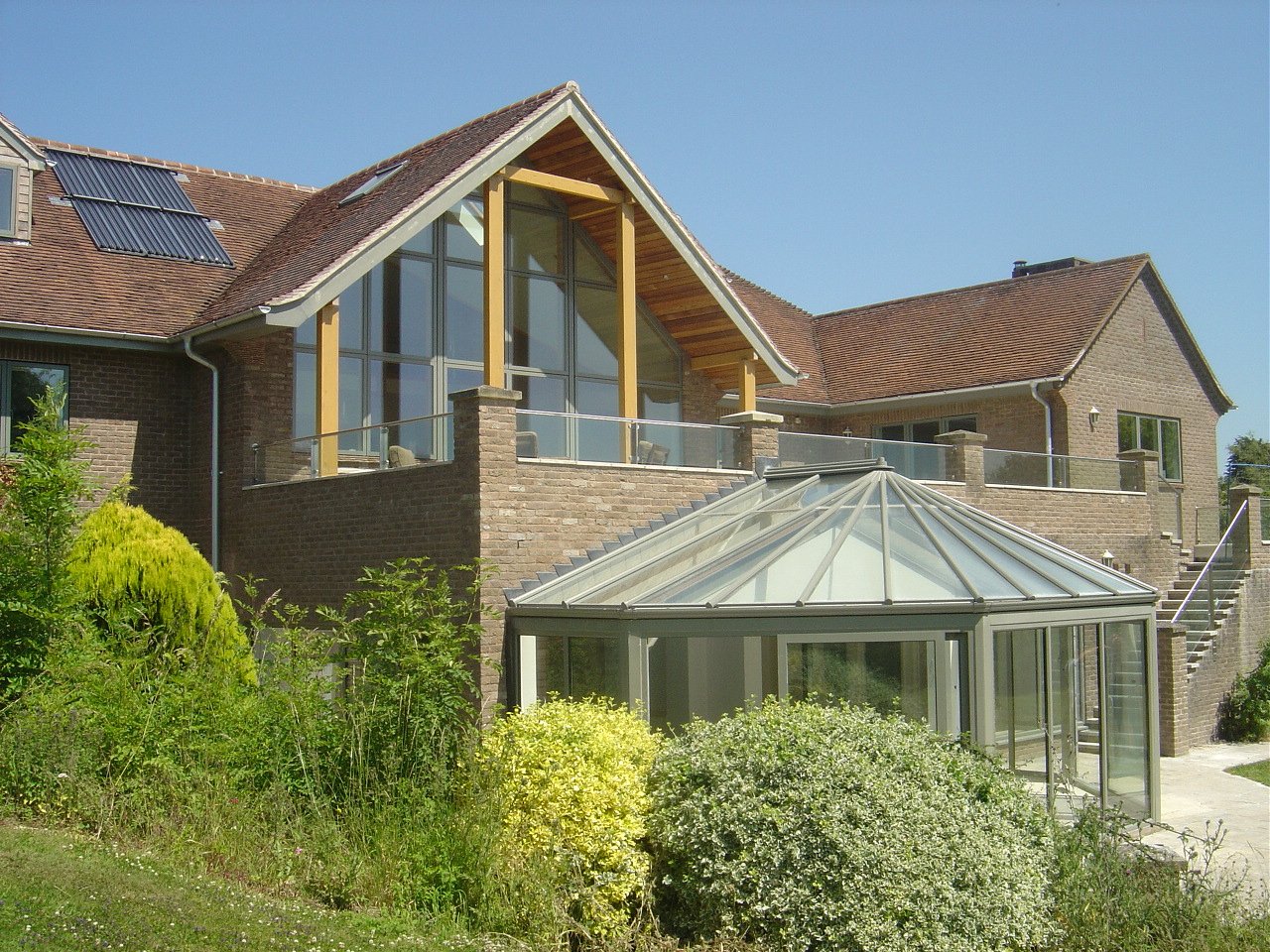 Modern house with brick exterior, large glass windows, a glass-enclosed sunroom, and a balcony on the upper level. The house has a sloped roof with solar panels and is surrounded by greenery.
