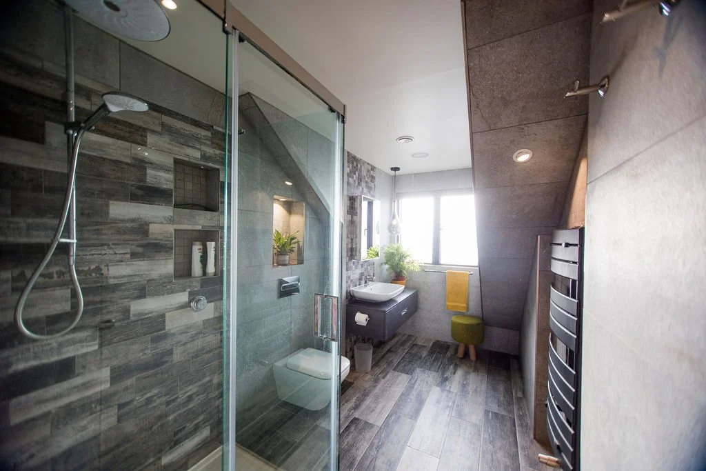 Modern bathroom featuring a walk-in shower with gray tiles, a wall-mounted sink, a window with natural light, a green stool, and a towel rack.
