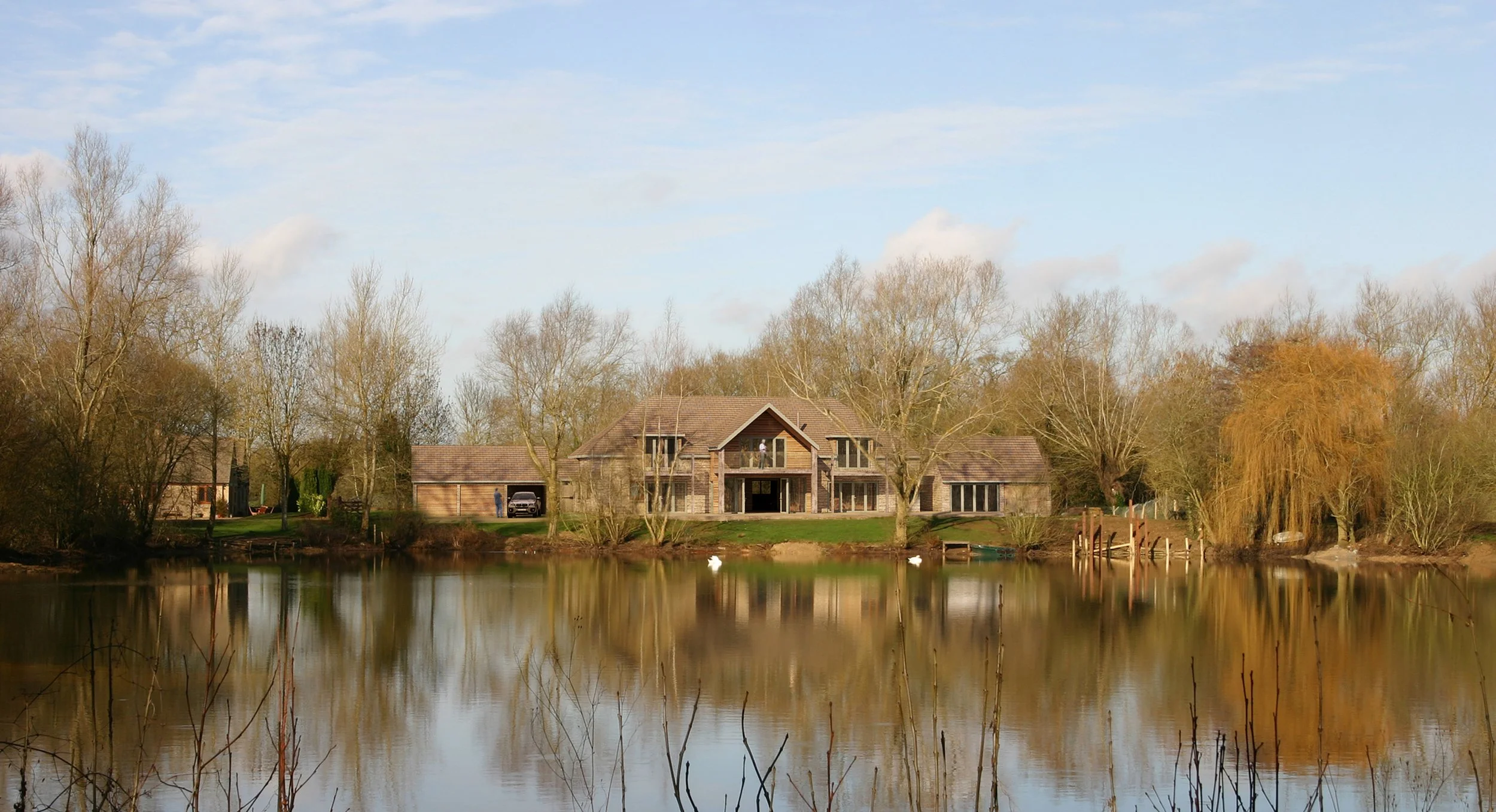 A house by a lake surrounded by leafless trees, with ducks swimming in the water, under a partly cloudy sky.