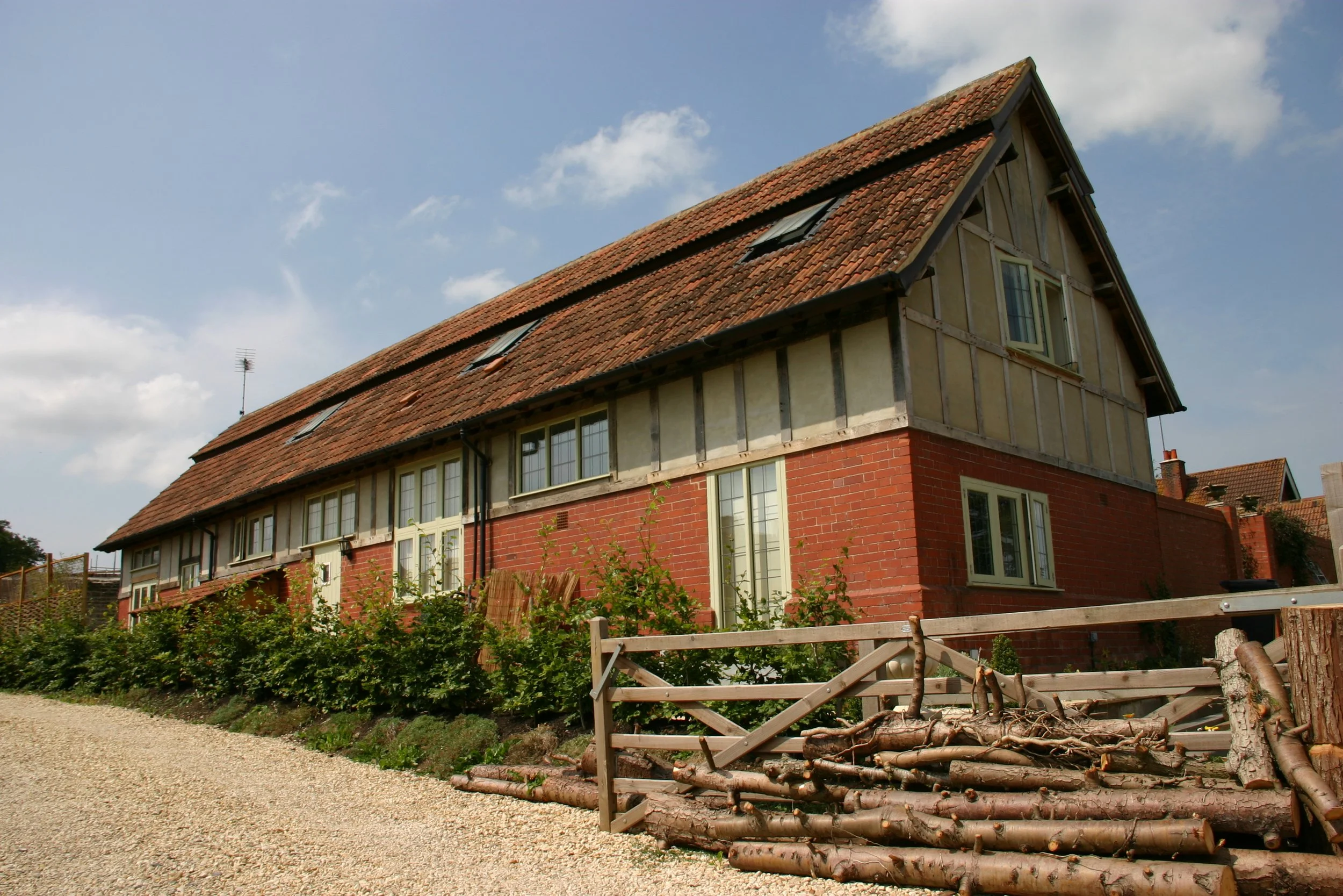 A house with brick and timber framing, a pitched roof with red tiles, multiple windows, and a small garden with bushes and logs in the foreground.