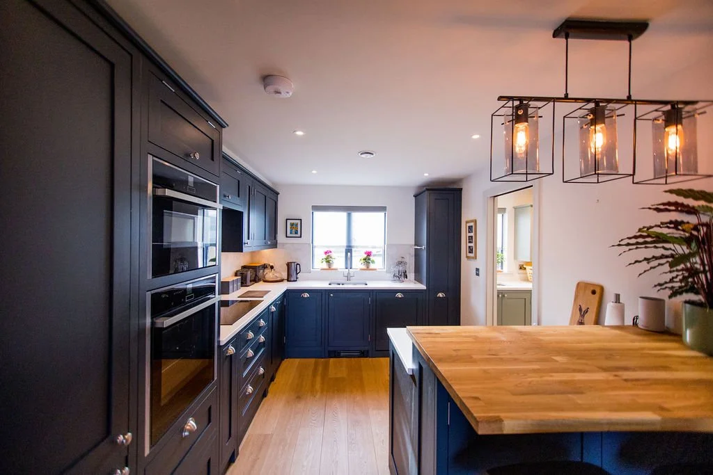 A modern kitchen with dark blue cabinets, a wooden island, and a window with potted plants, illuminated by pendant lights.