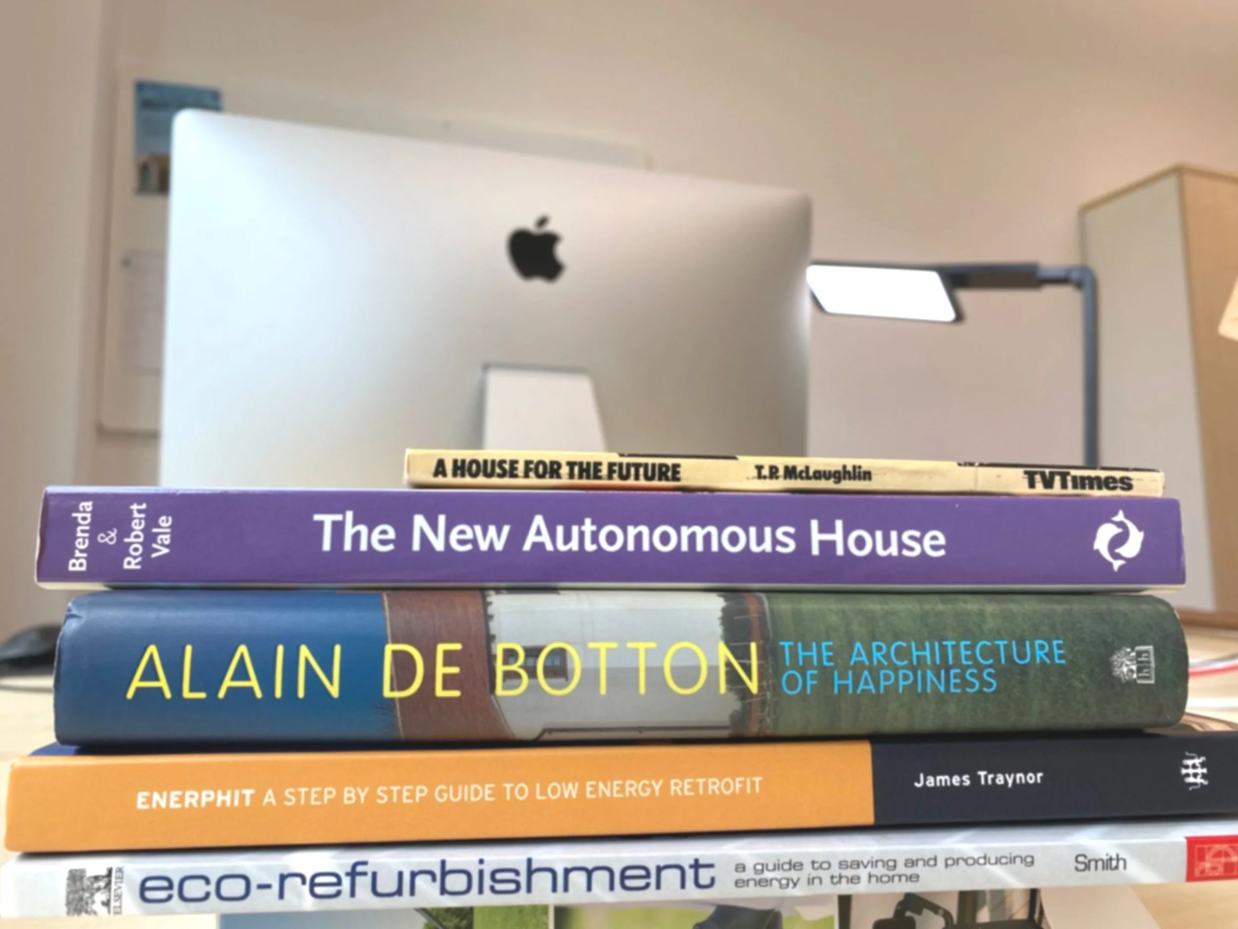 A stack of five books on a desk with an iMac computer in the background. The book titles are 'A House for the Future', 'The New Autonomous House', 'The Architecture of Happiness', 'Enerphit', and 'Eco-refurbishment'.
