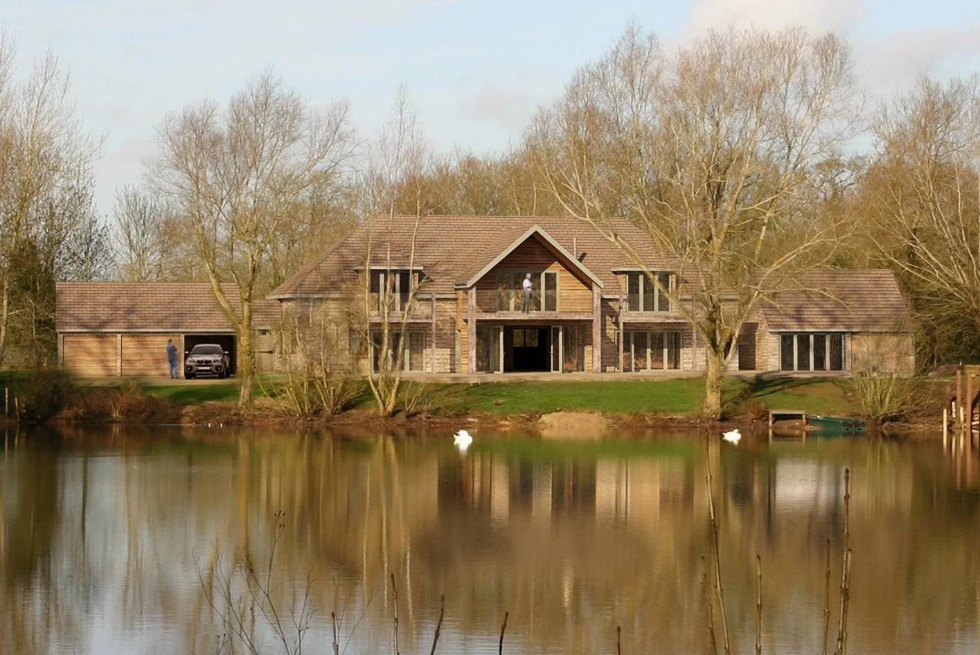 Large house with a brick and wood exterior, situated by a lake with ducks, surrounded by leafless trees, and a person standing in the driveway.