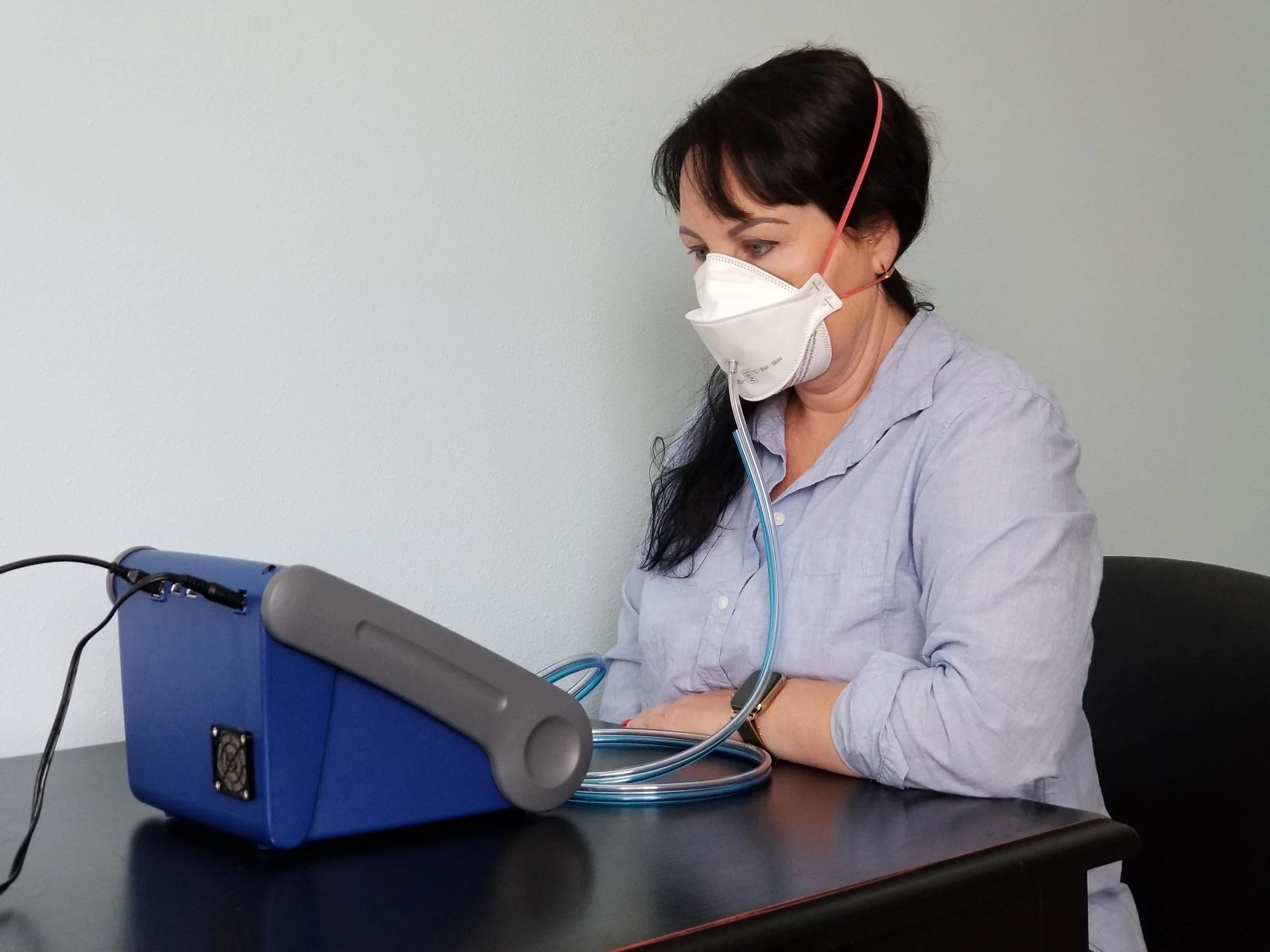 A woman wearing a face mask and a light gray shirt measures her oxygen levels using a pulse oximeter connected to a blue medical device on a black table.