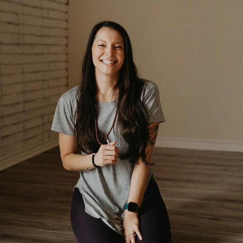 A woman with long dark hair smiling while sitting on the floor in a room with wooden flooring and a brick wall background.