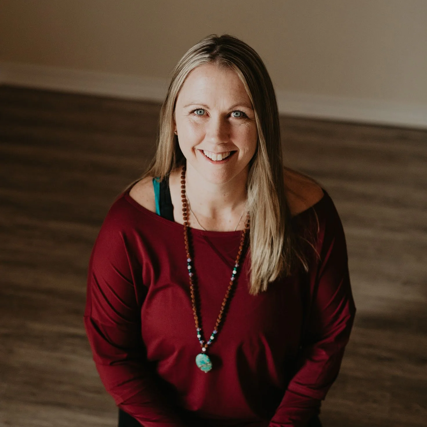 A woman with long blonde hair, blue eyes, wearing a maroon top, layered necklaces, and smiling.